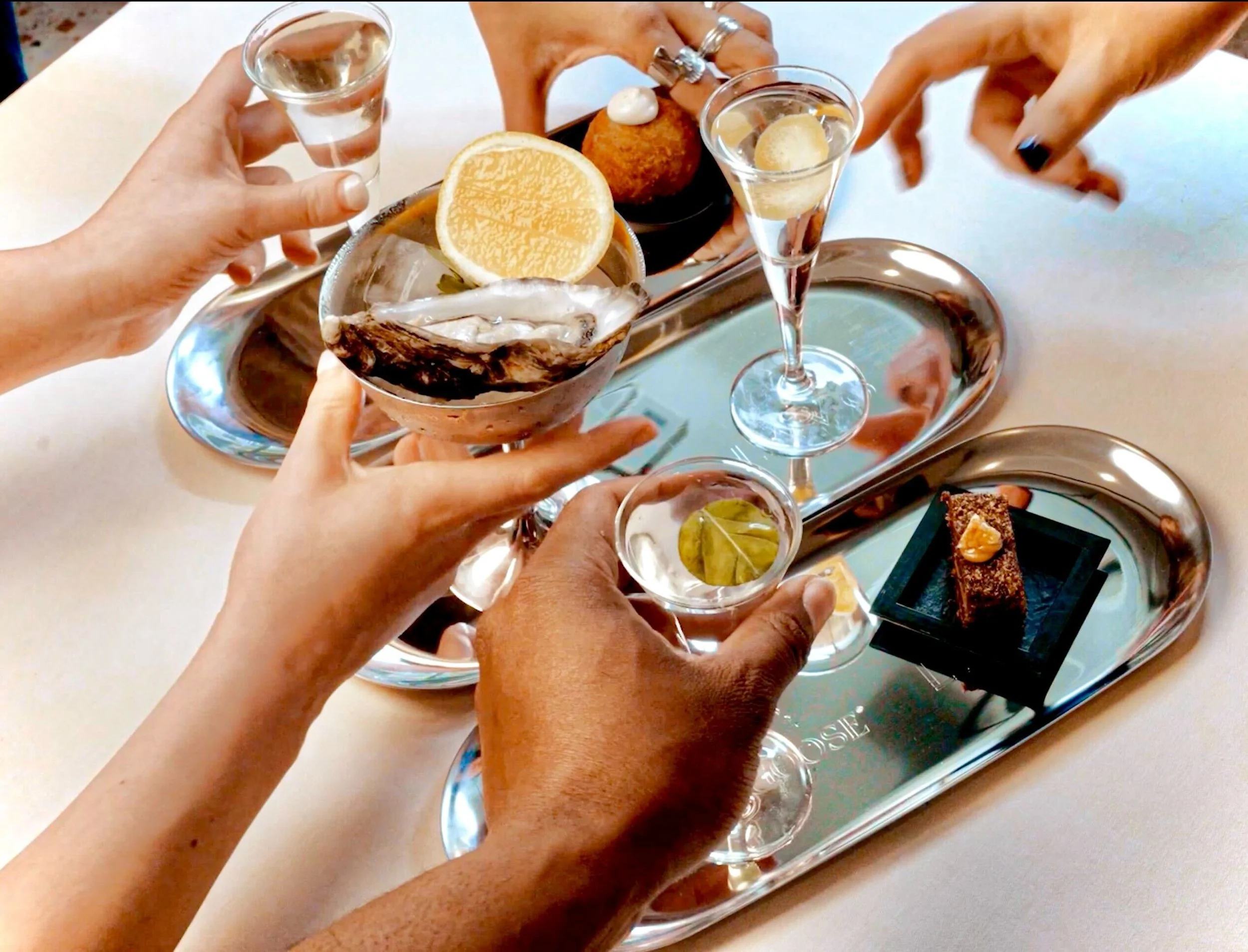 People toasting with shot glasses at a silver tray with seafood, lemon slices, and desserts.