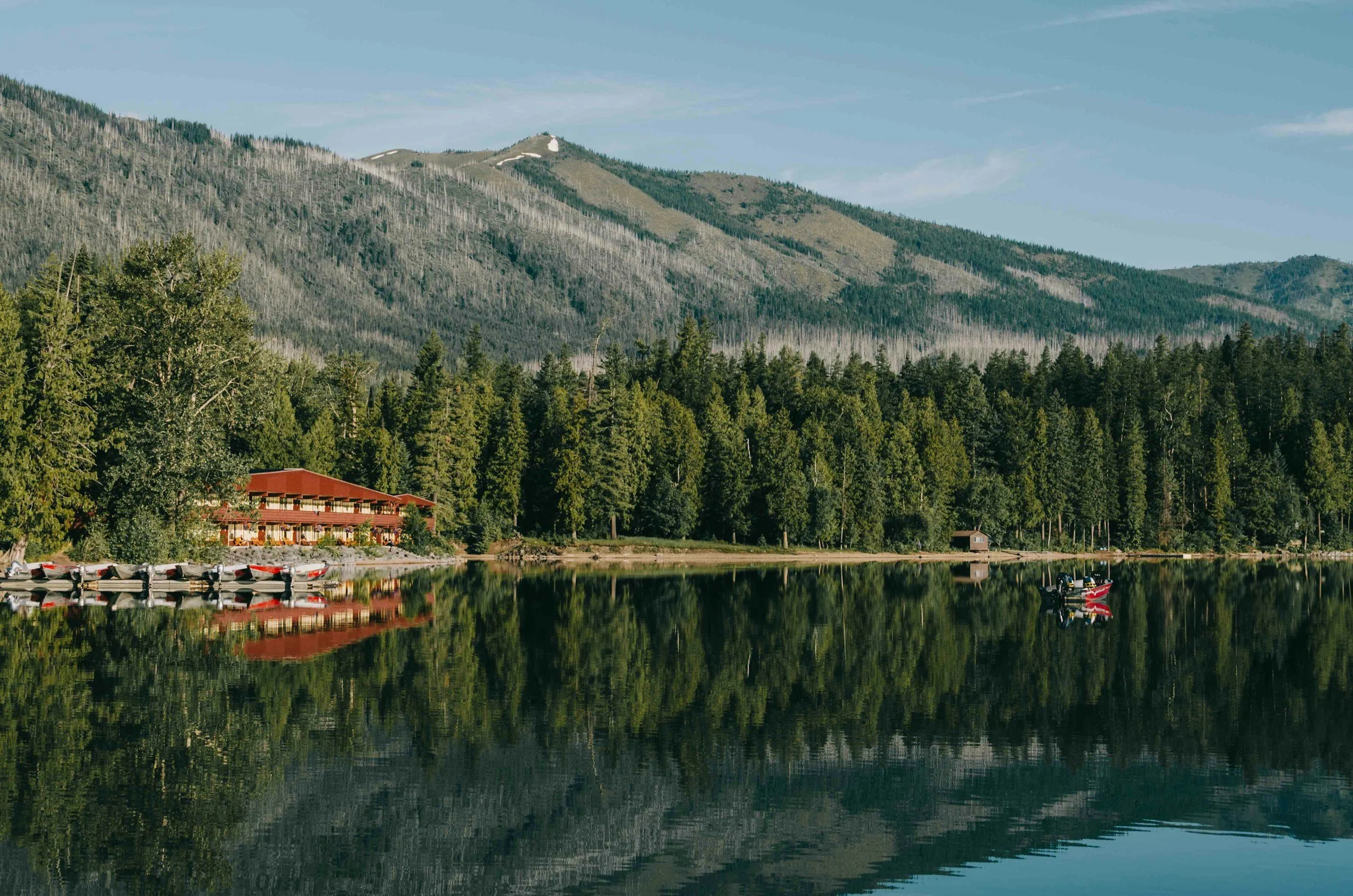 A serene lake reflecting a dense forest and mountains in the background, with a red building and boats docked along the shoreline.