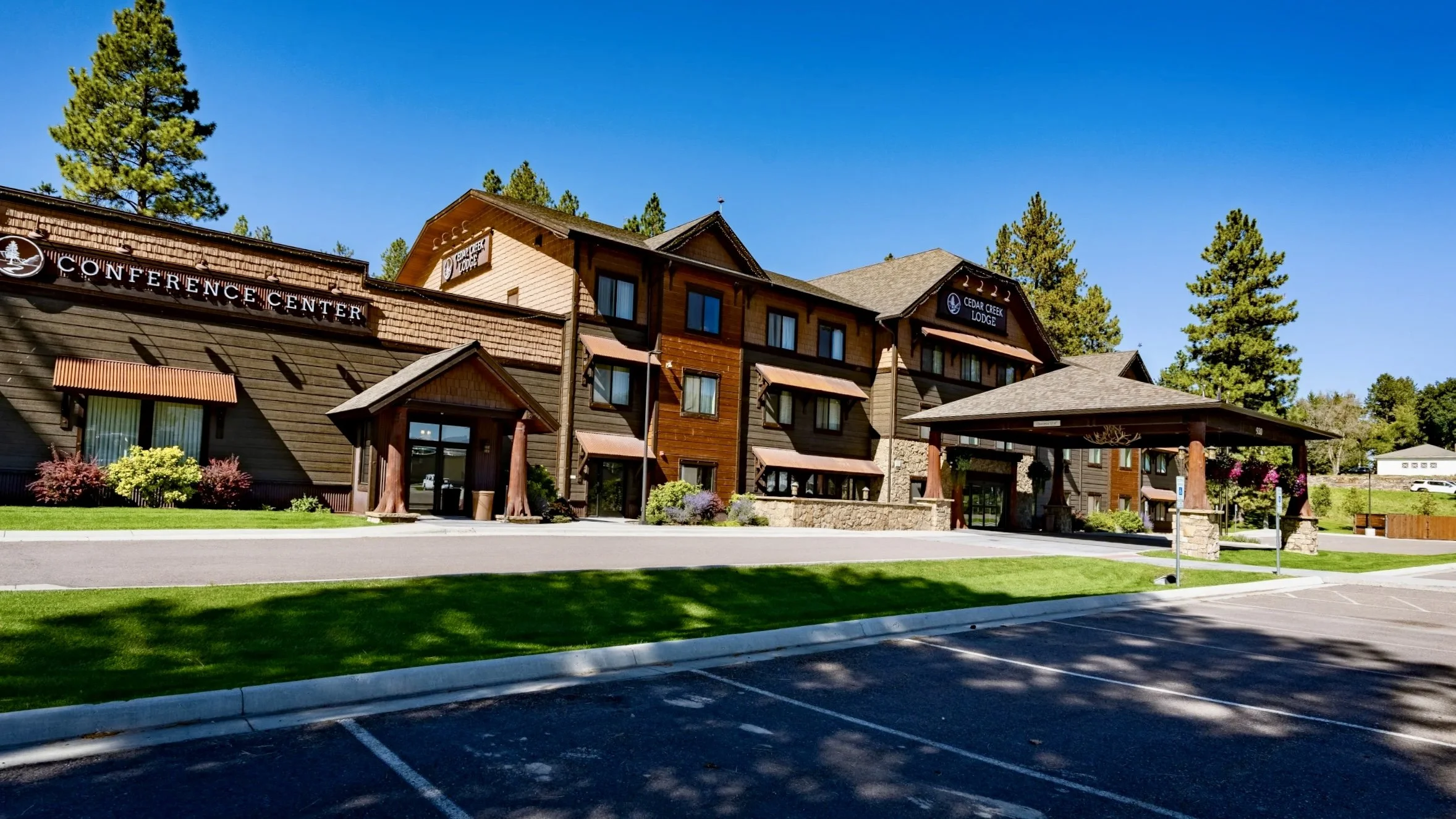 Exterior view of Cedar Creek Lodge, a multi-story building with wooden siding, surrounded by green lawns, shrubs, and trees under a clear blue sky. The building has multiple windows with awnings, a conference center sign, and a covered entrance.