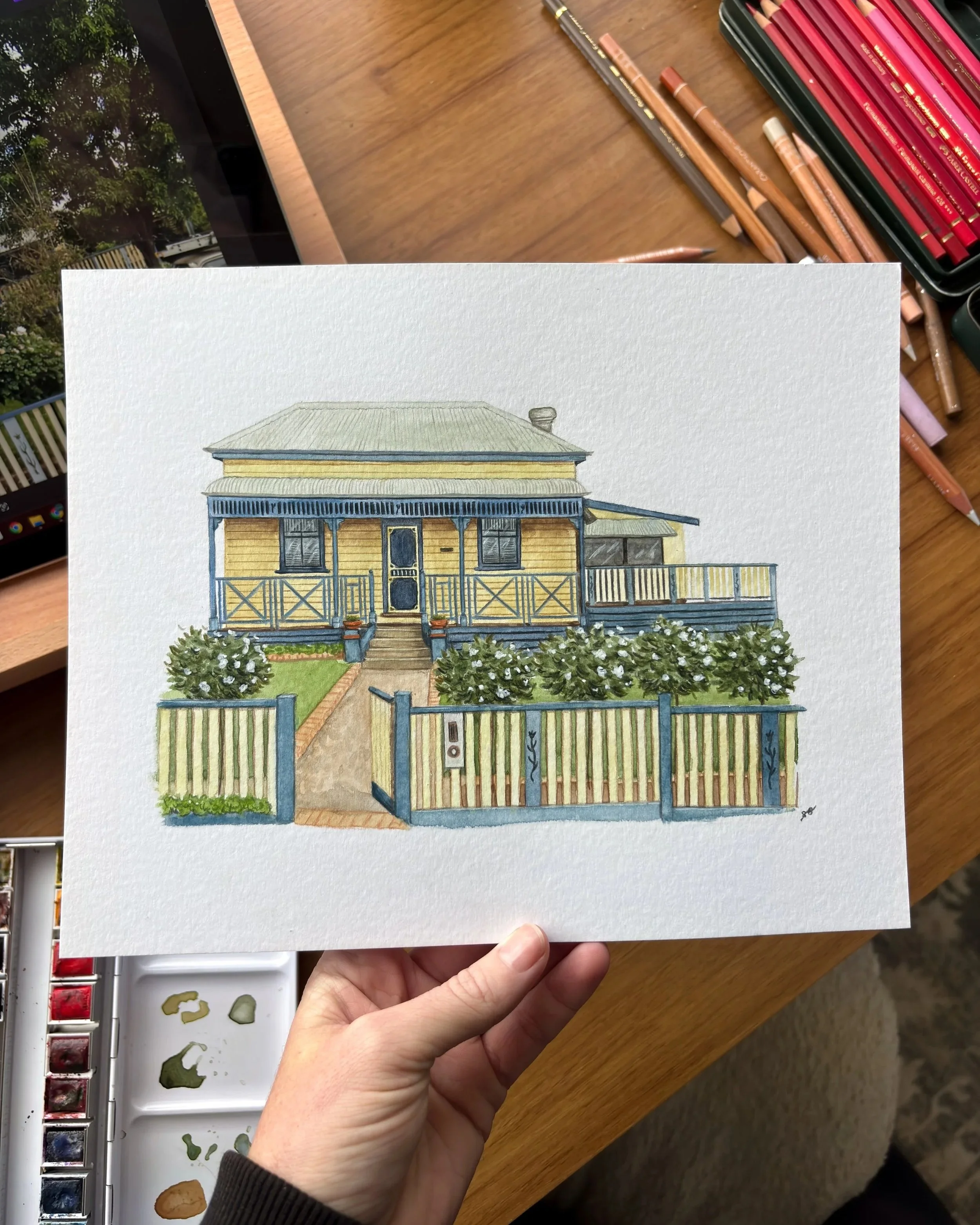 A detailed architectural watercolor painting of a yellow, wooden, two-story house with a porch, staircase, and a gated front yard, held in a person's hand above a wooden table with a watercolor palette and art supplies.