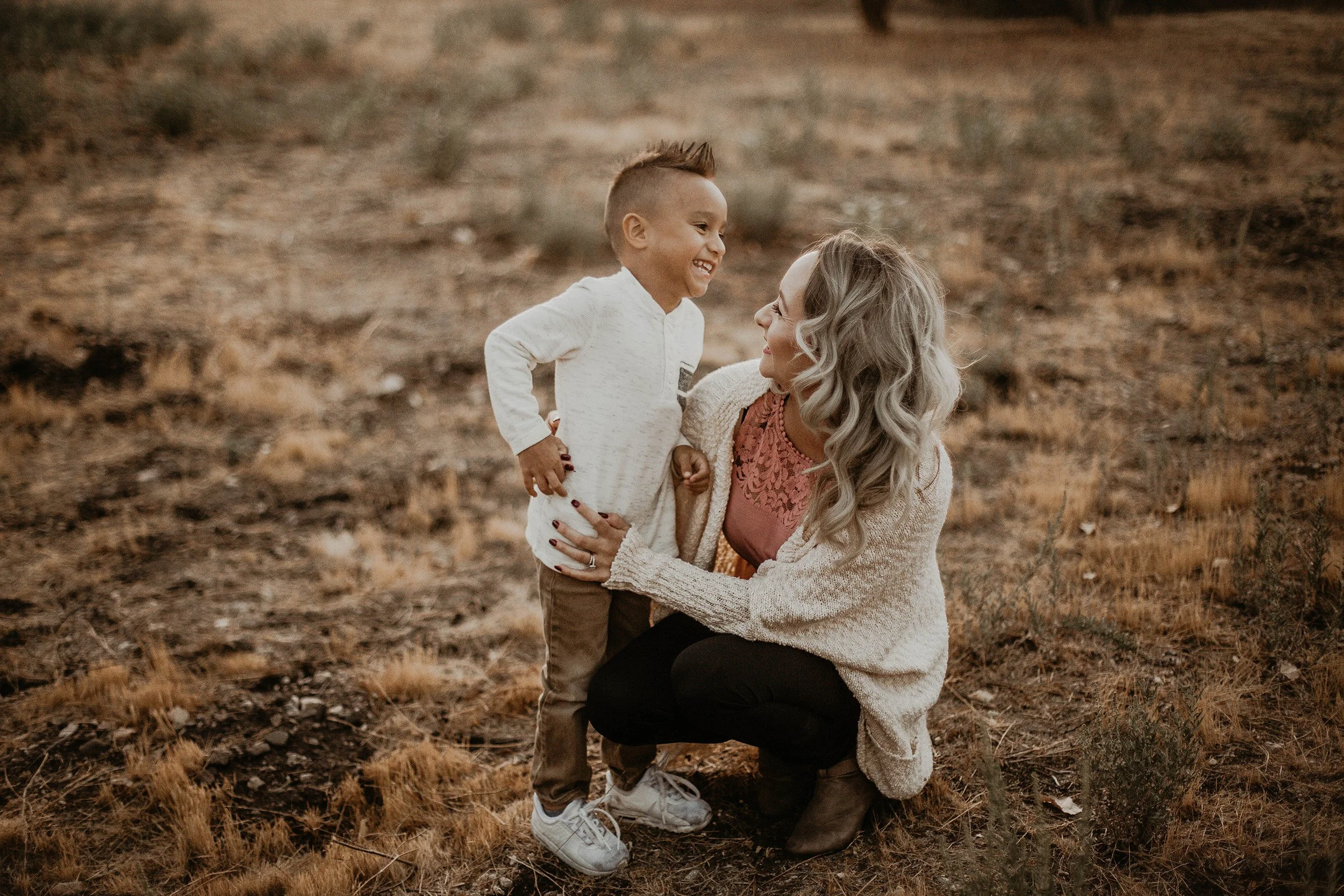 A woman with blonde curly hair kneels on the ground, smiling at a young boy with a short haircut, as they hold hands and look at each other in a field with dry grass.