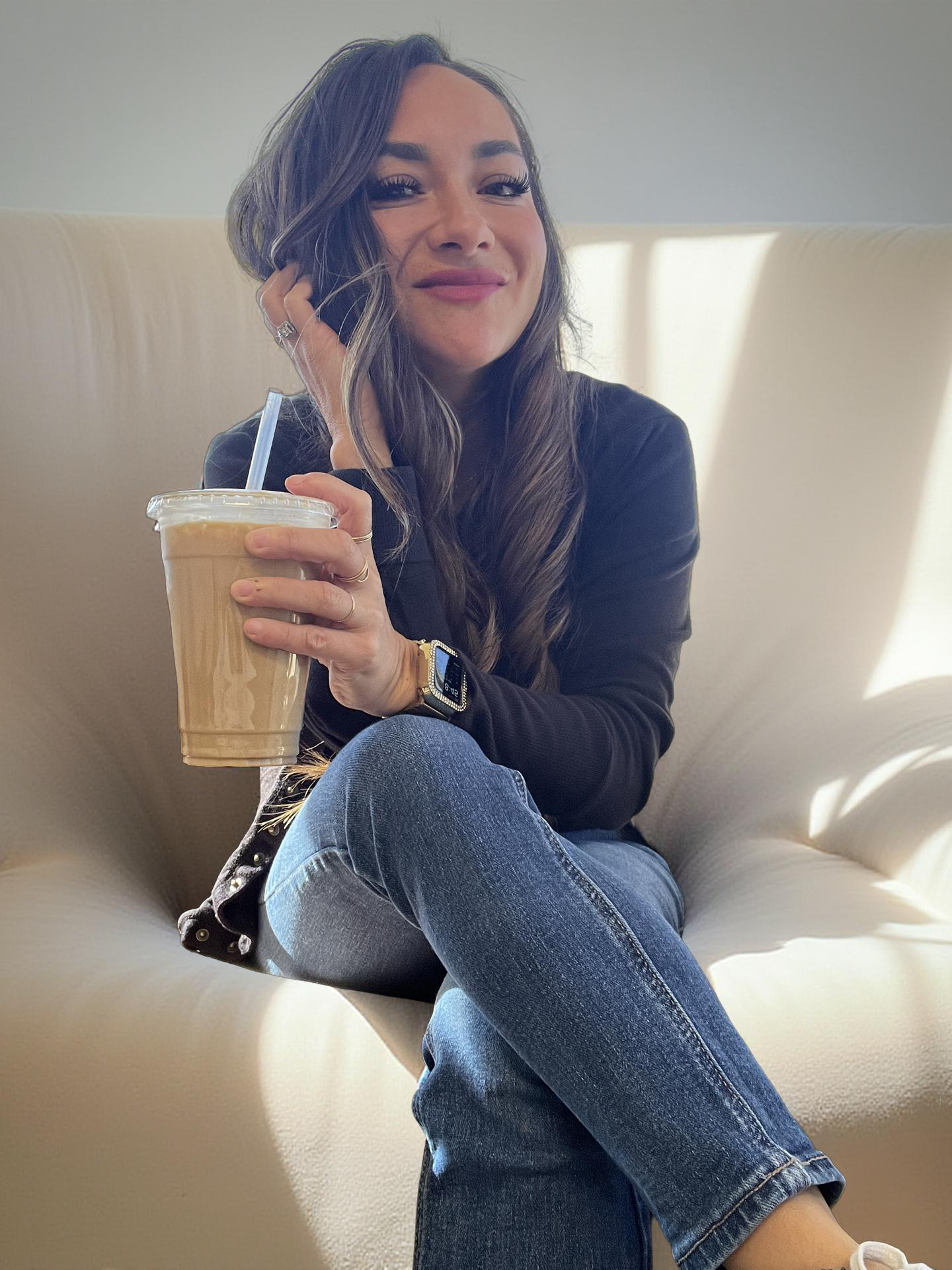 A young woman with long brown hair, wearing a black jacket and blue jeans, sitting on a cream-colored sofa, holding a large iced coffee, smiling at the camera.