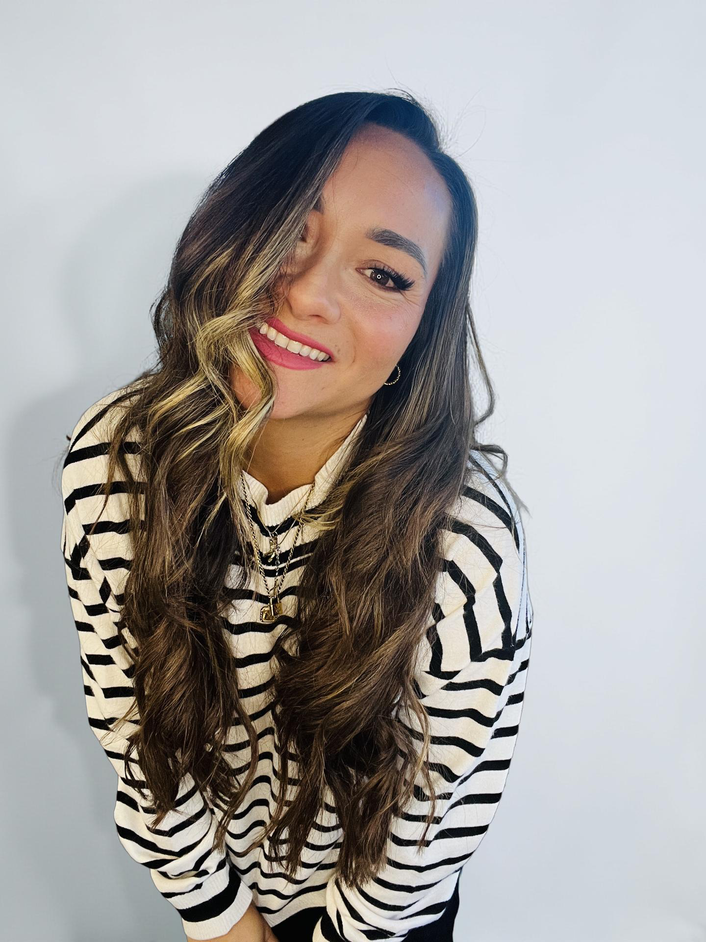 A young woman with long wavy hair, smiling, wearing a black and white striped shirt and layered necklaces, standing against a plain white wall.