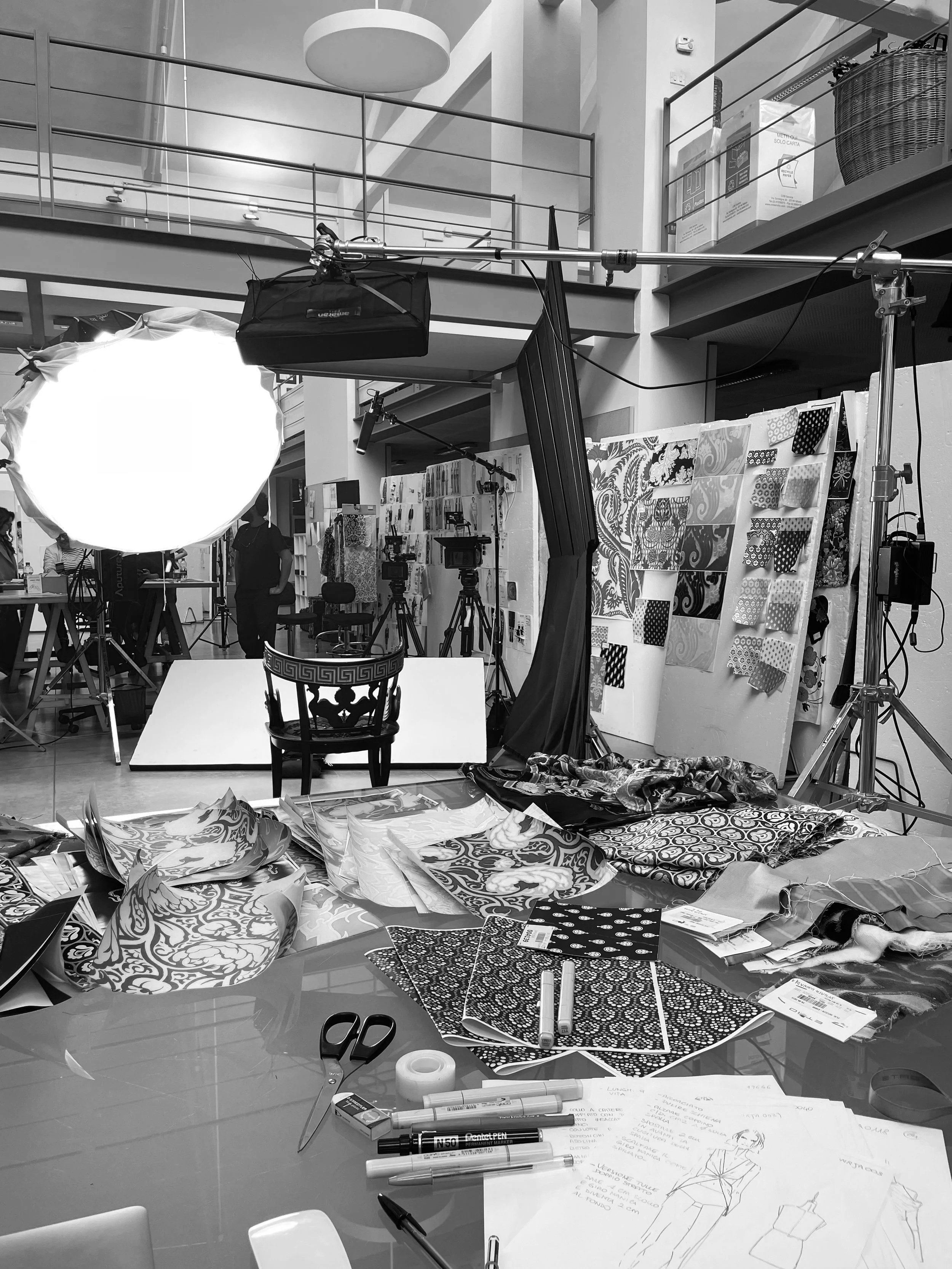 Indoor photo of a fashion or textile design studio with fabrics, sketches, and lighting equipment. People and camera setups are visible in the background.