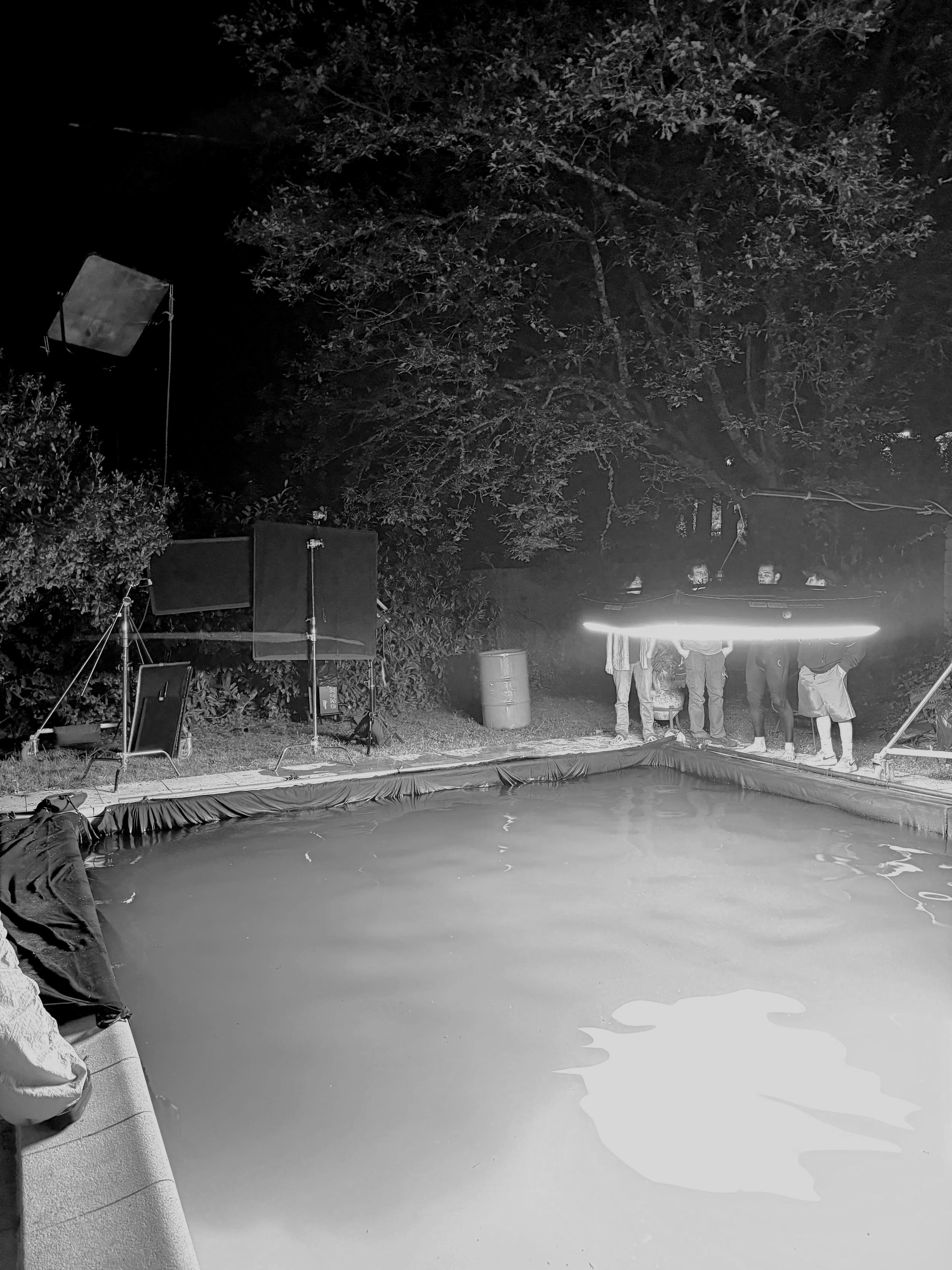 A group of people standing near the edge of a swimming pool at night, with film lighting equipment set up around it, and trees in the background.