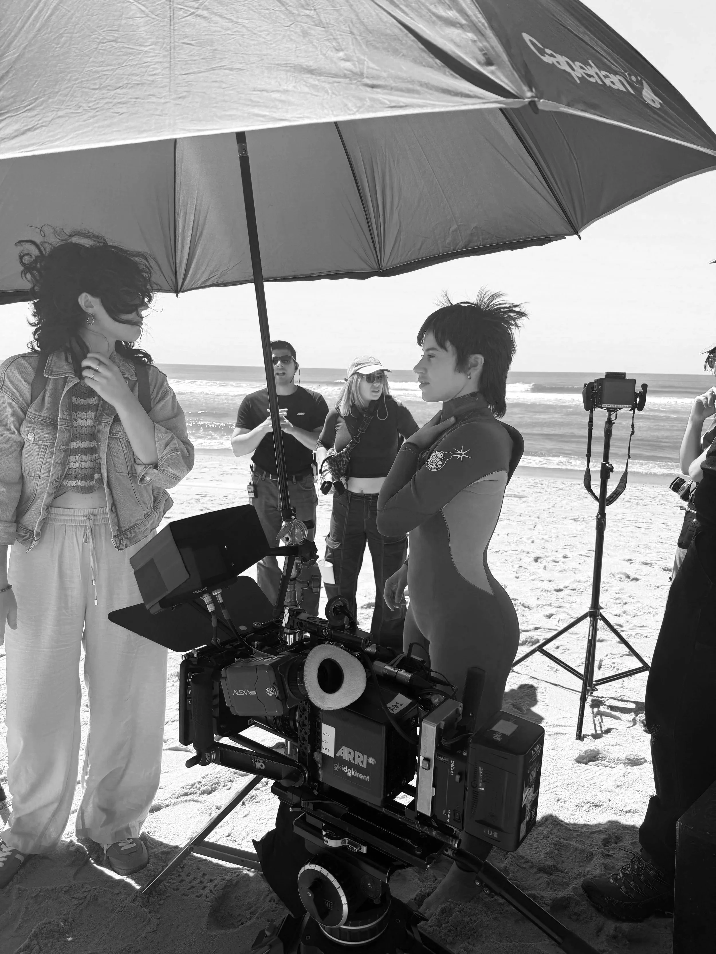 A film crew on a beach under a large umbrella filming a woman in a wetsuit. The crew includes camera operators and assistants, with equipment set up around them.