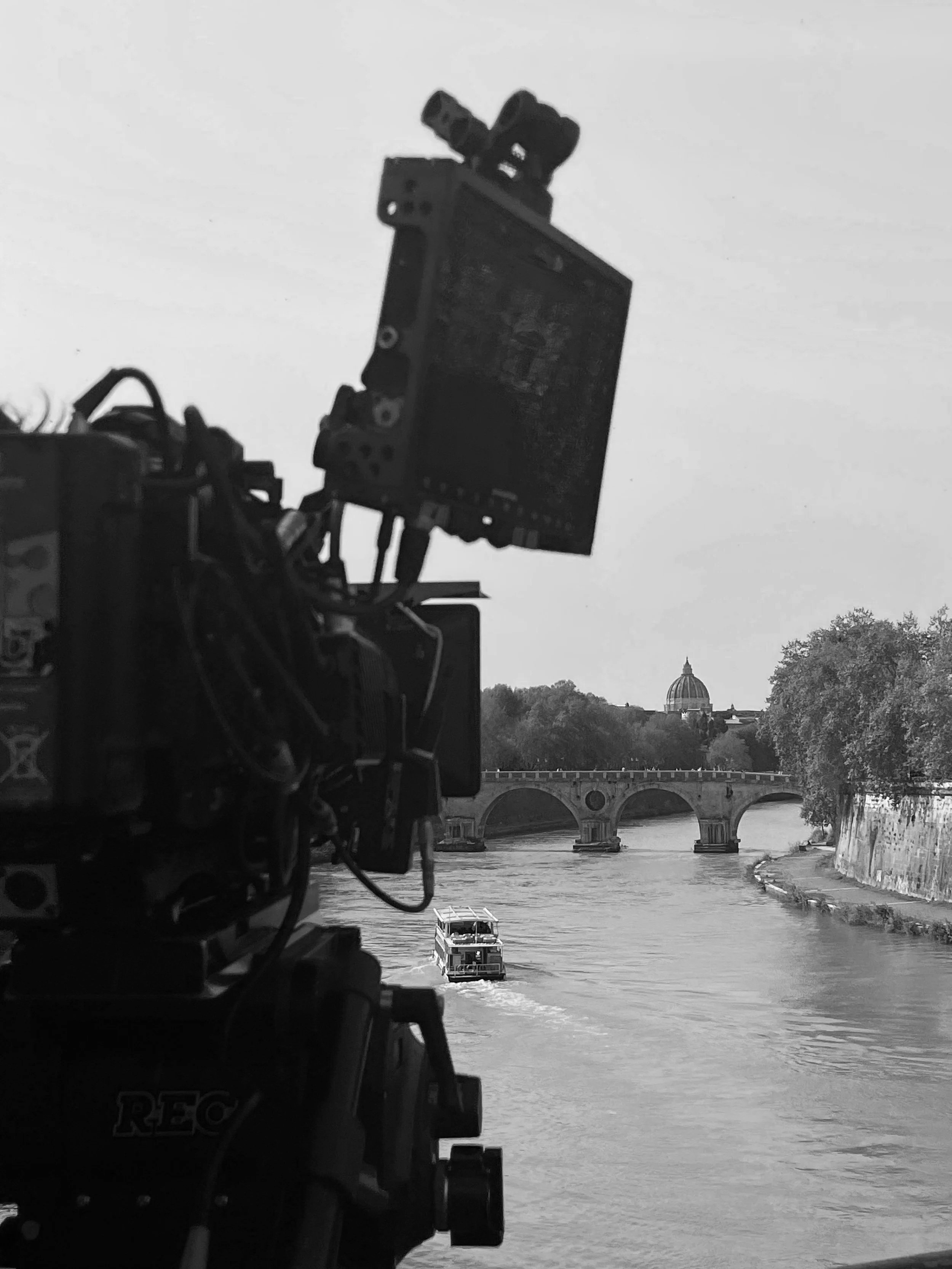 Film camera setup on a riverbank capturing a boat on the river, with a bridge and a domed building in the background.