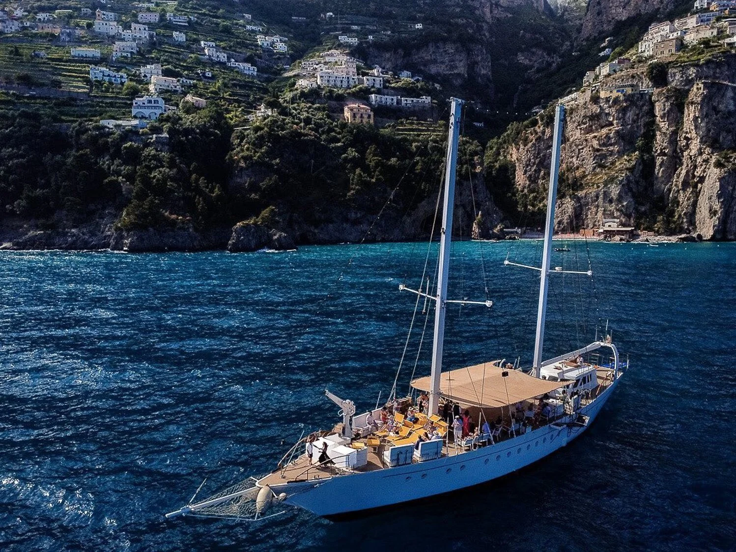 A sailboat with a beige canopy floating on dark blue water near a rocky coastline with white buildings on terraced green hillside.