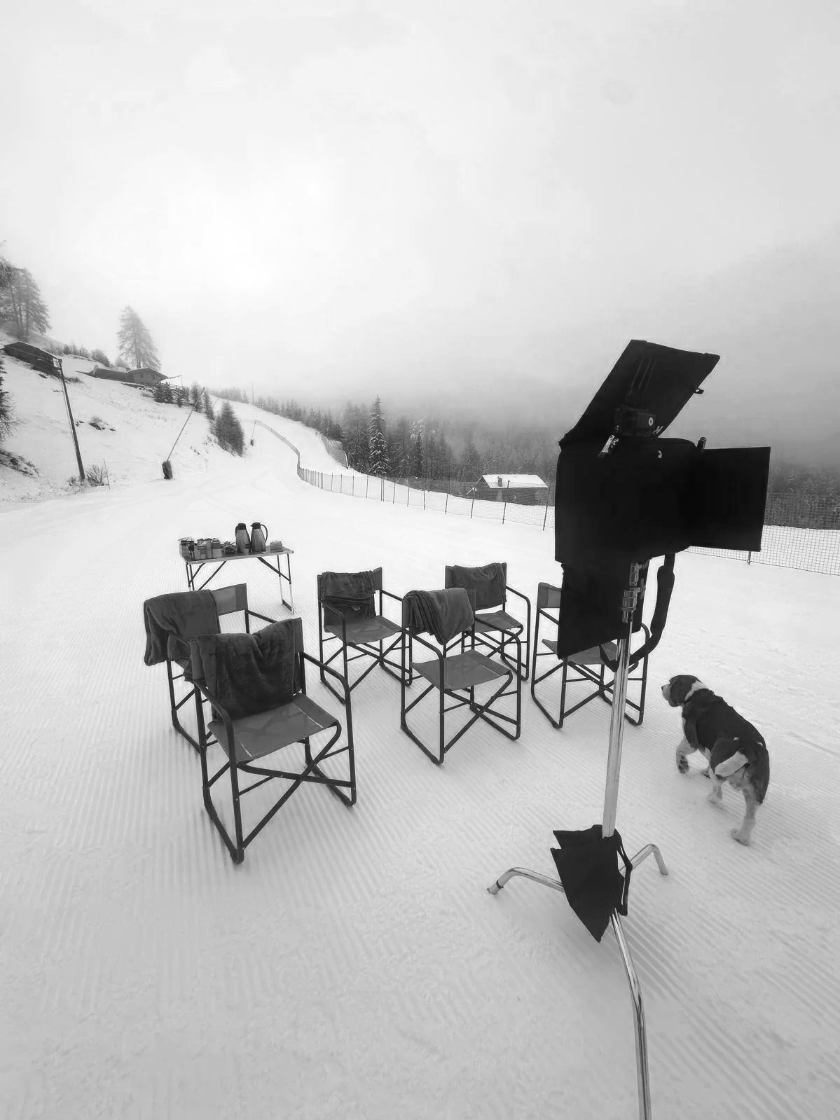 An outdoor film shoot set in a snowy, mountainous landscape with six chairs, a table with coffee pots, a film light, and a dog nearby.