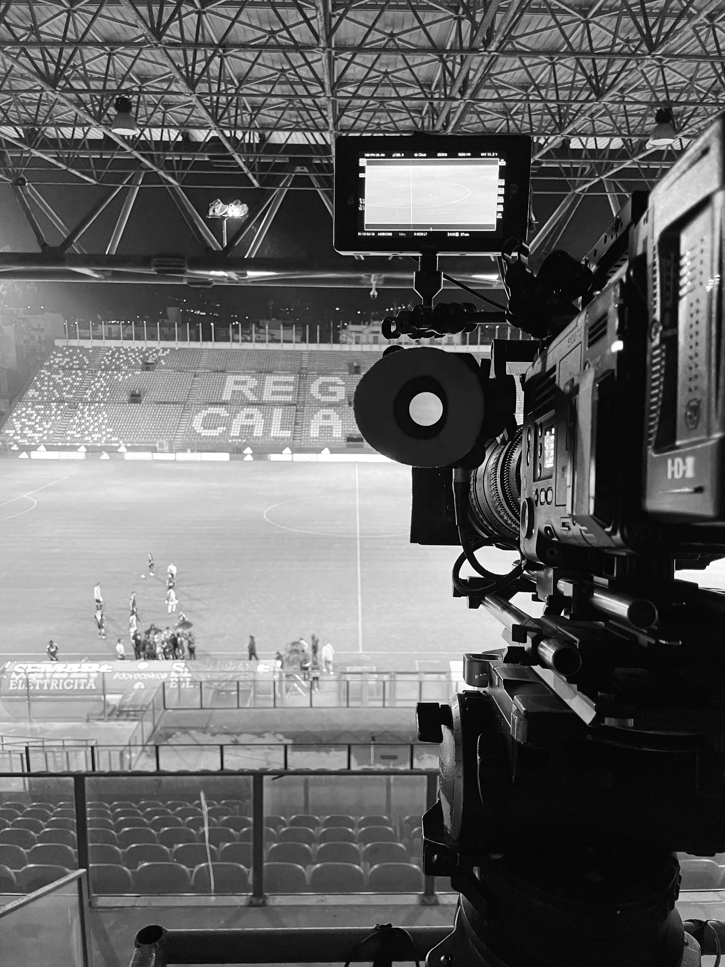 View of a soccer stadium field from behind the goal, with a professional camera on a tripod capturing a game, and empty seats in the foreground.