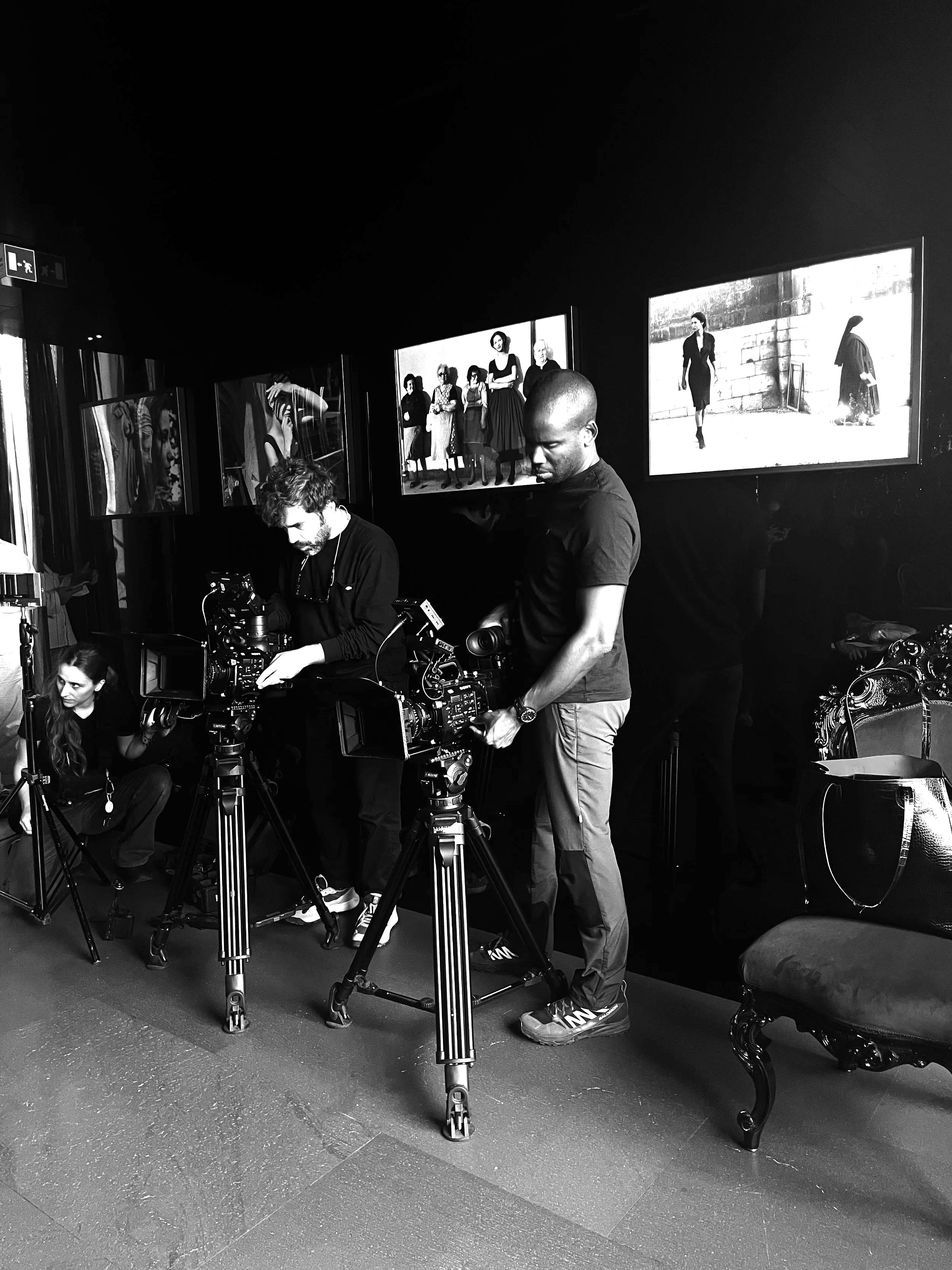 Photographers setting up cameras in front of framed photographs on a dark wall in an art gallery or exhibit.