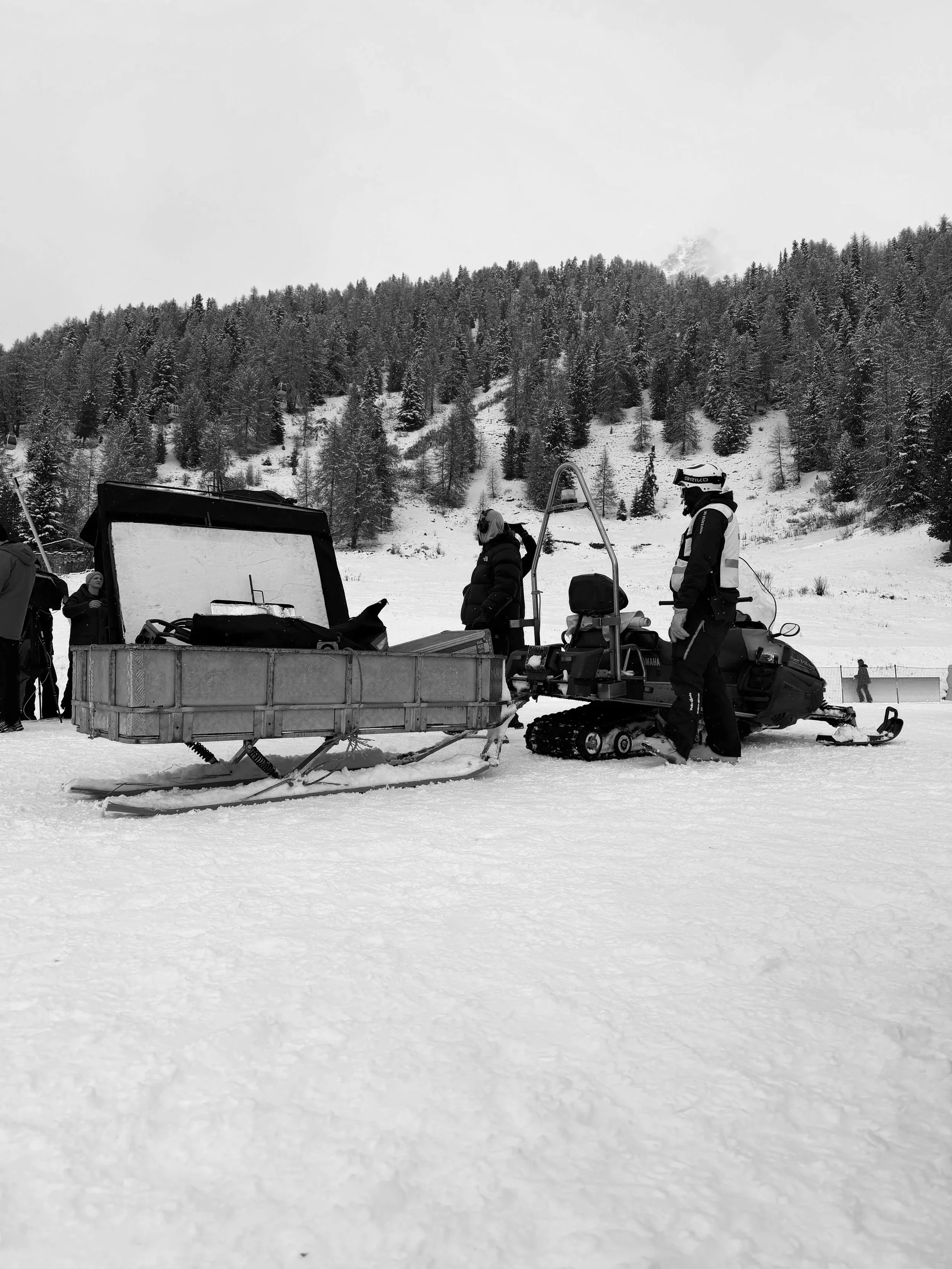 People on a snowmobile trailer in a snowy landscape with trees and hills in the background.