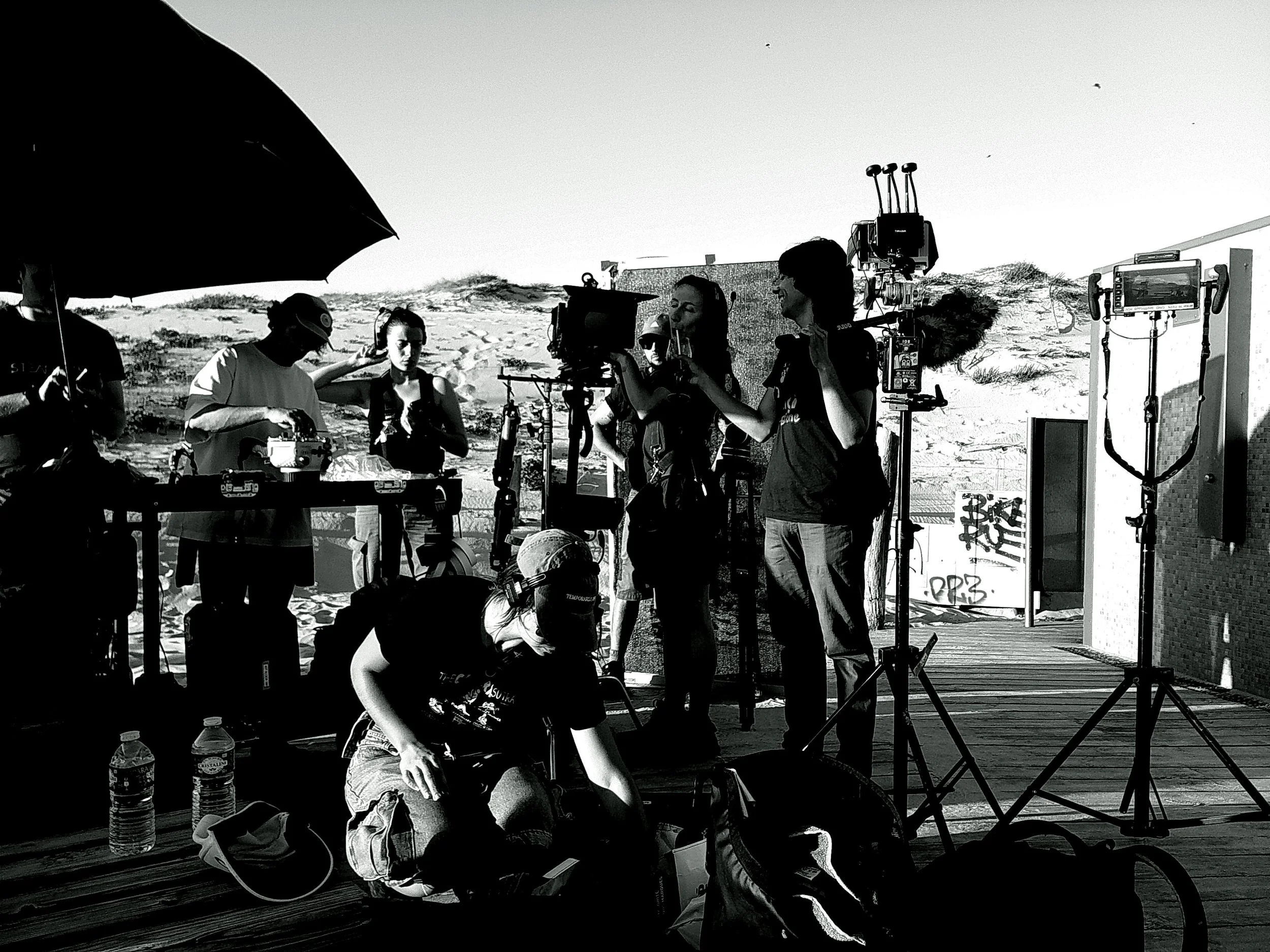 Filming crew setting up equipment on a beach with sandy dunes in the background.