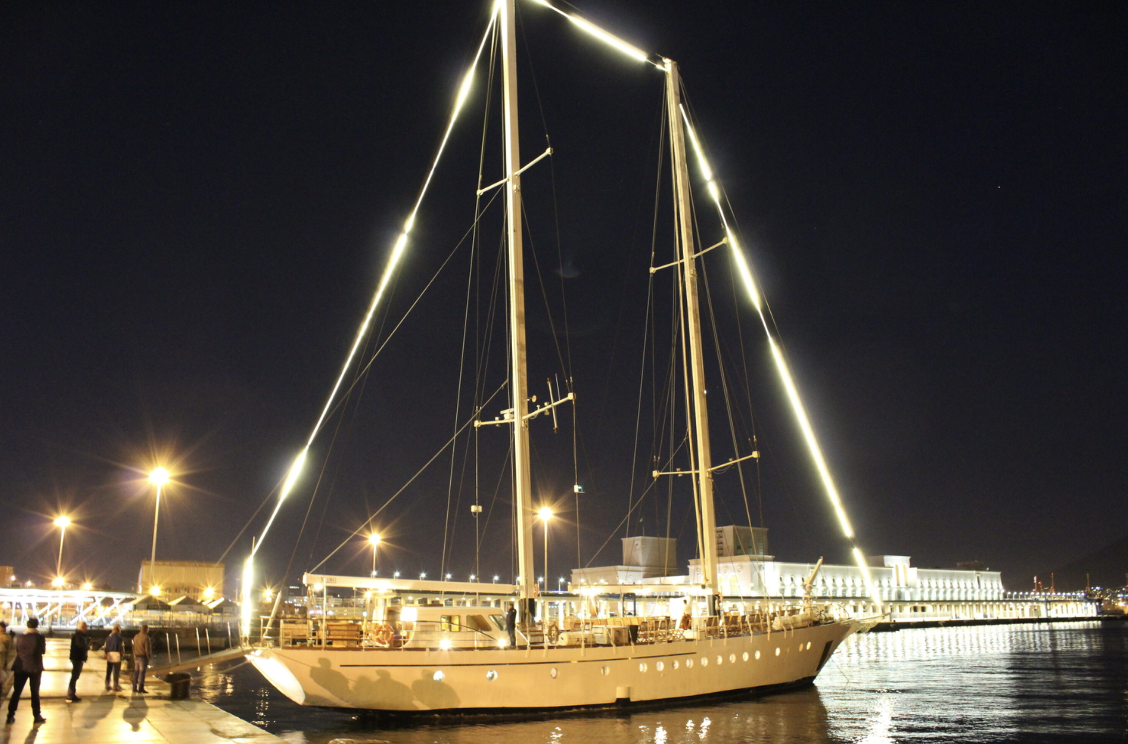 A large sailboat docked at a pier at night with bright lights illuminating it and the surrounding area.
