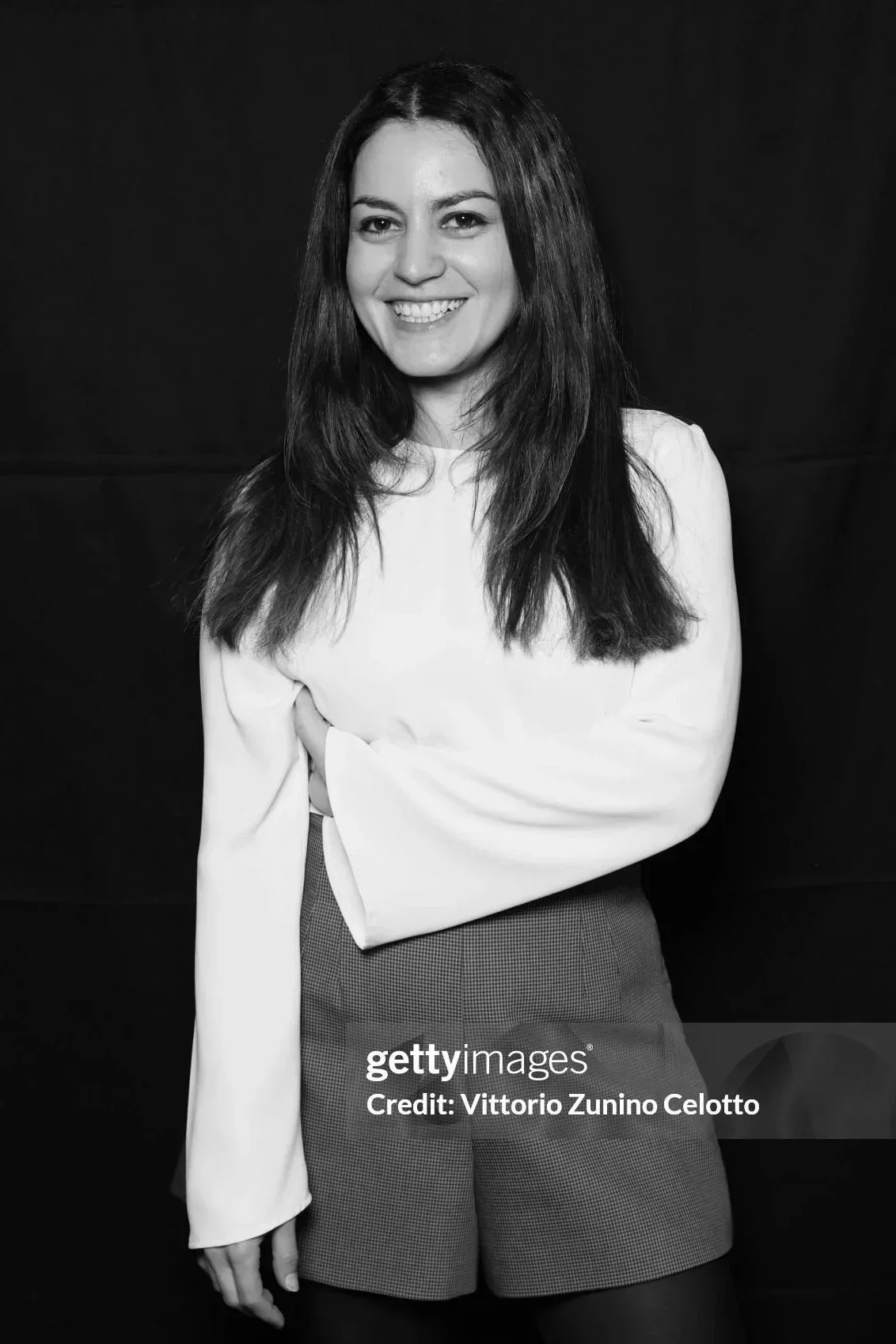 Black and white portrait of a woman with long dark hair, smiling, wearing a white long-sleeve top and checkered shorts, standing against a dark background.