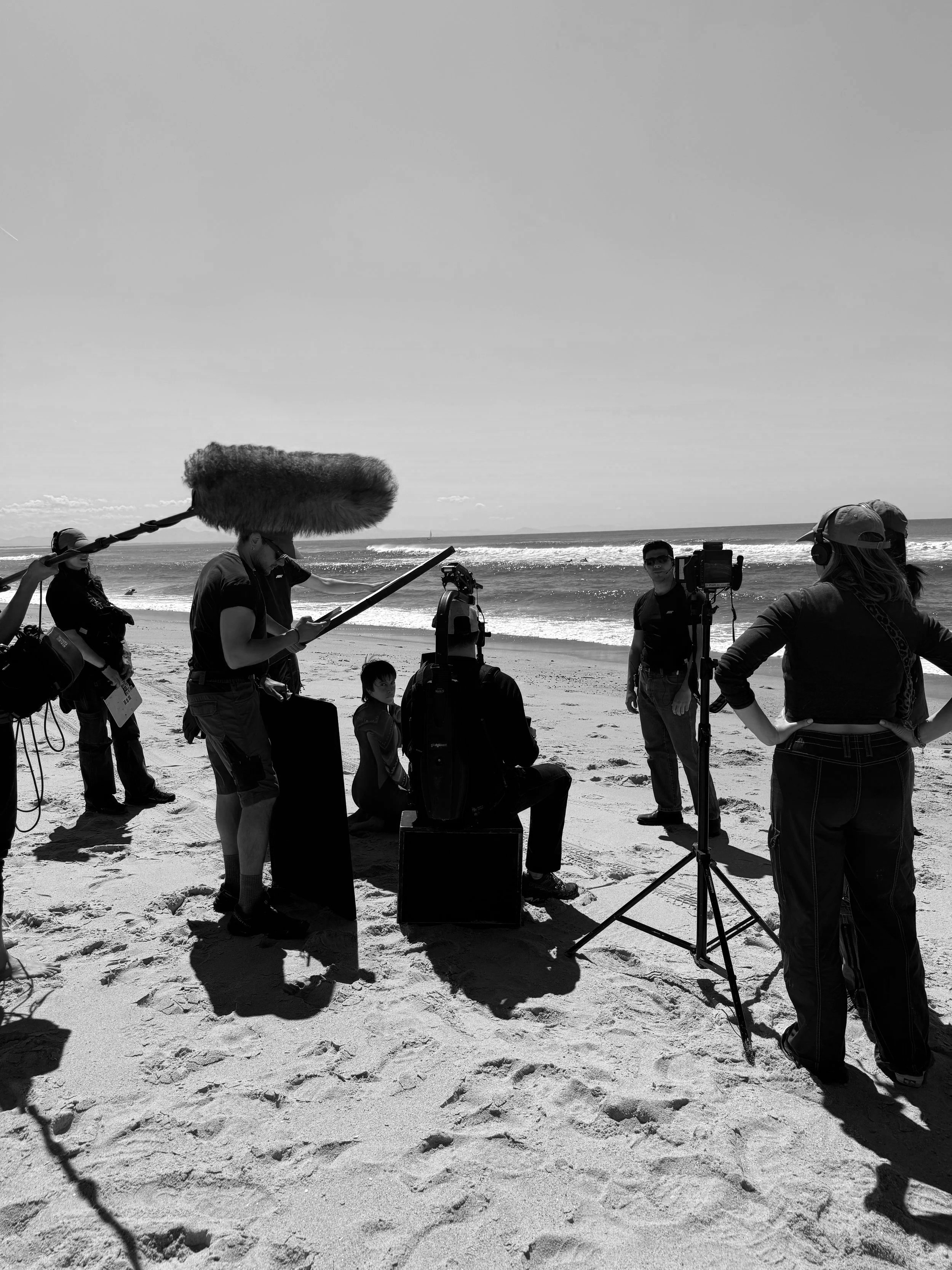 Filming crew on a beach with a seated person in front of a camera, surrounded by microphones and equipment, with the ocean in the background.