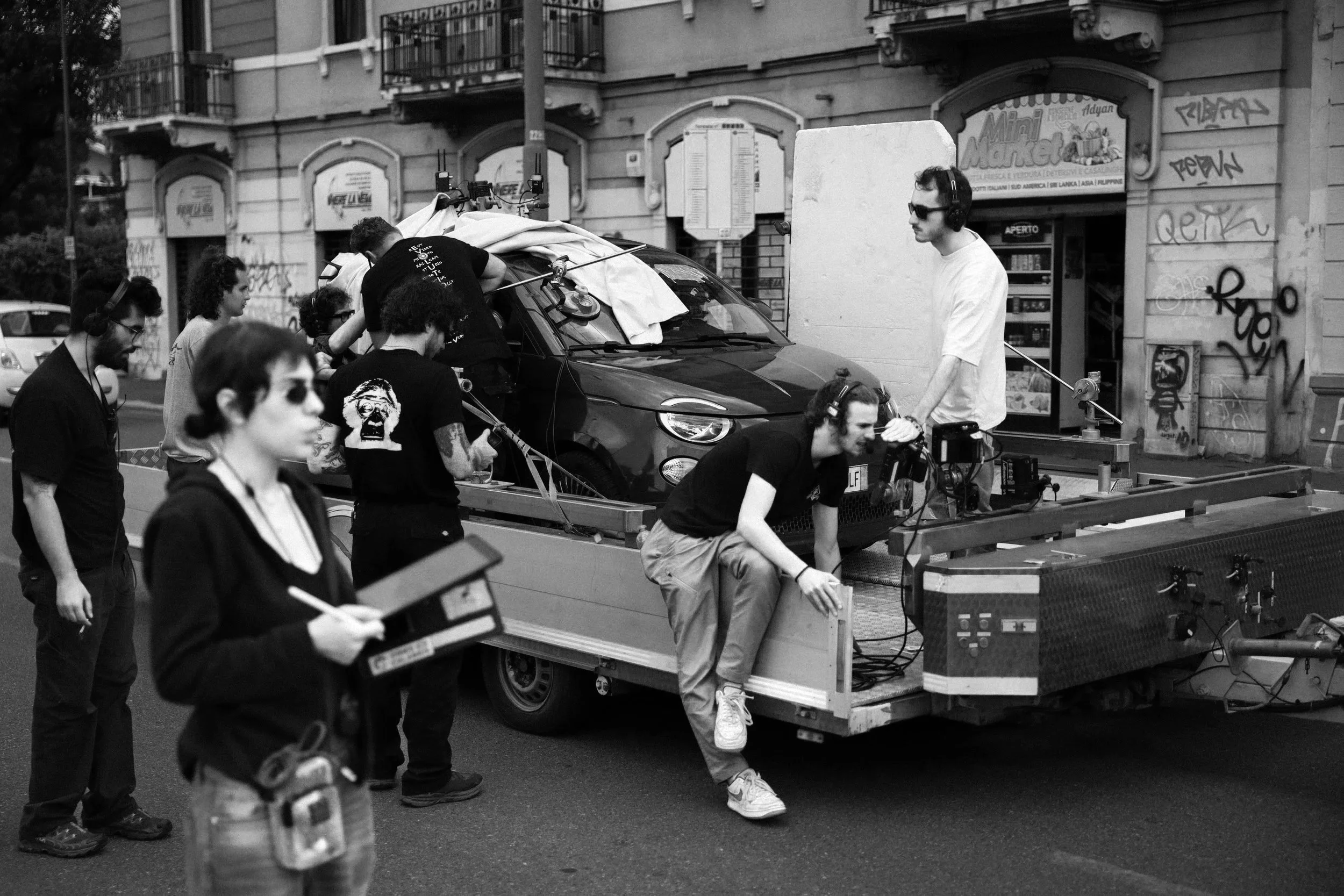 Black and white photo of a film crew filming a car stunt on a city street. Several crew members are around a car mounted on a trailer, with one person in white wearing headphones operating equipment, and others taking notes or observing. The setting appears urban with graffiti on the walls.