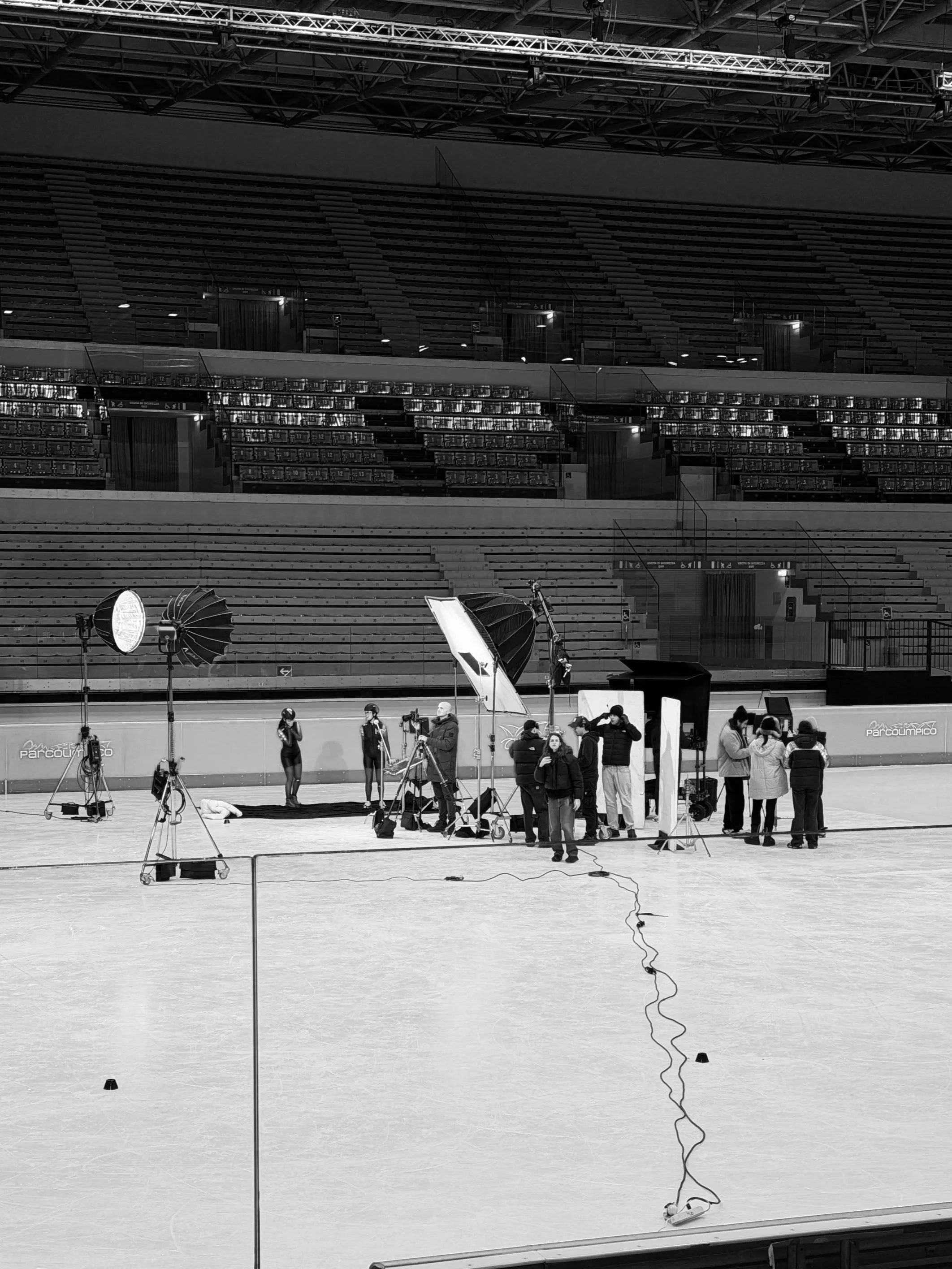 A filming crew setting up equipment on an indoor ice rink for a photoshoot or video shoot, with large studio lights, umbrellas, cameras, and a backdrop.