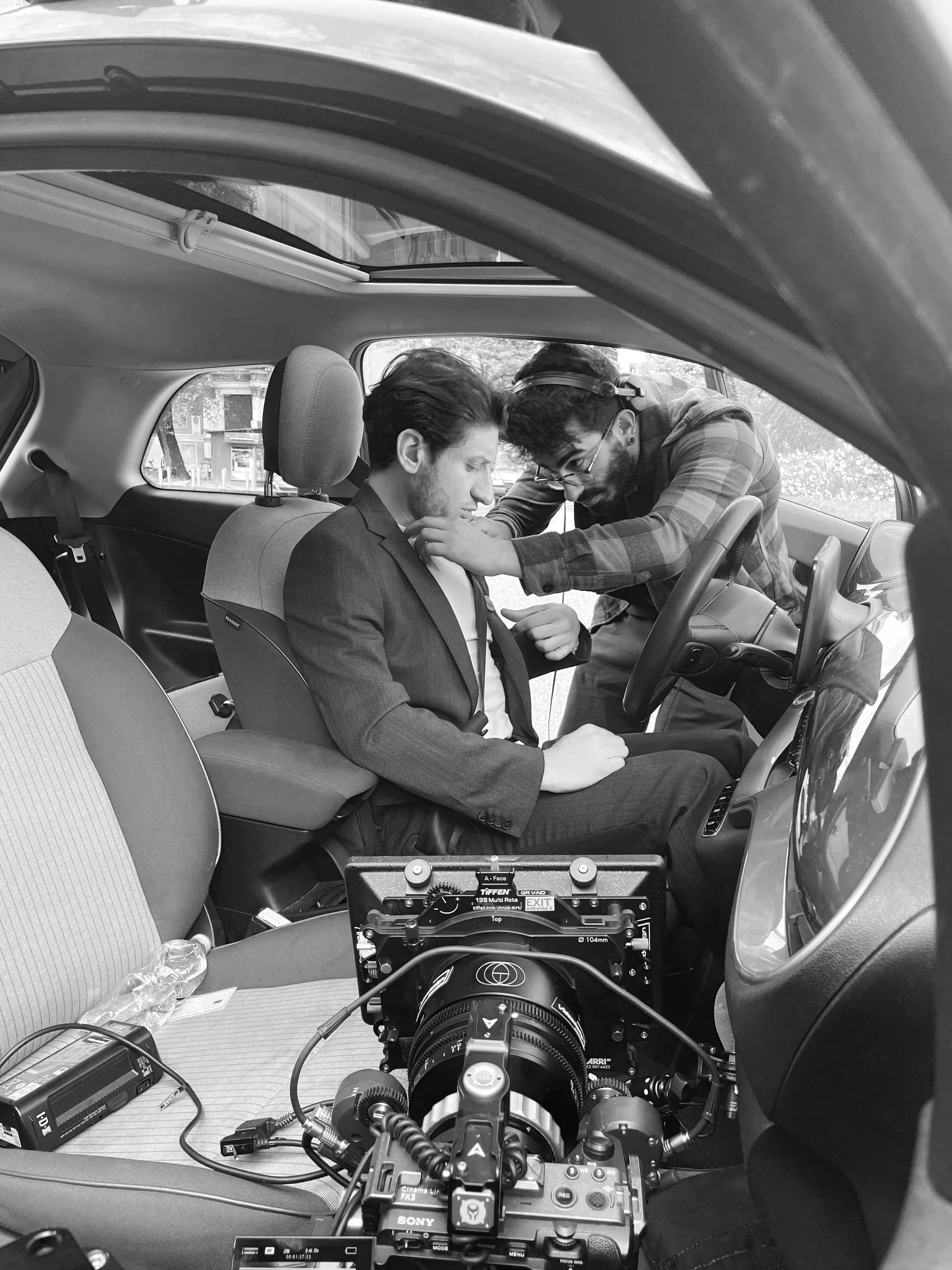 A man in a suit sitting in the driver's seat of a car being prepared for filming or photography by a crew member adjusting his shirt, with camera equipment set up inside the car.