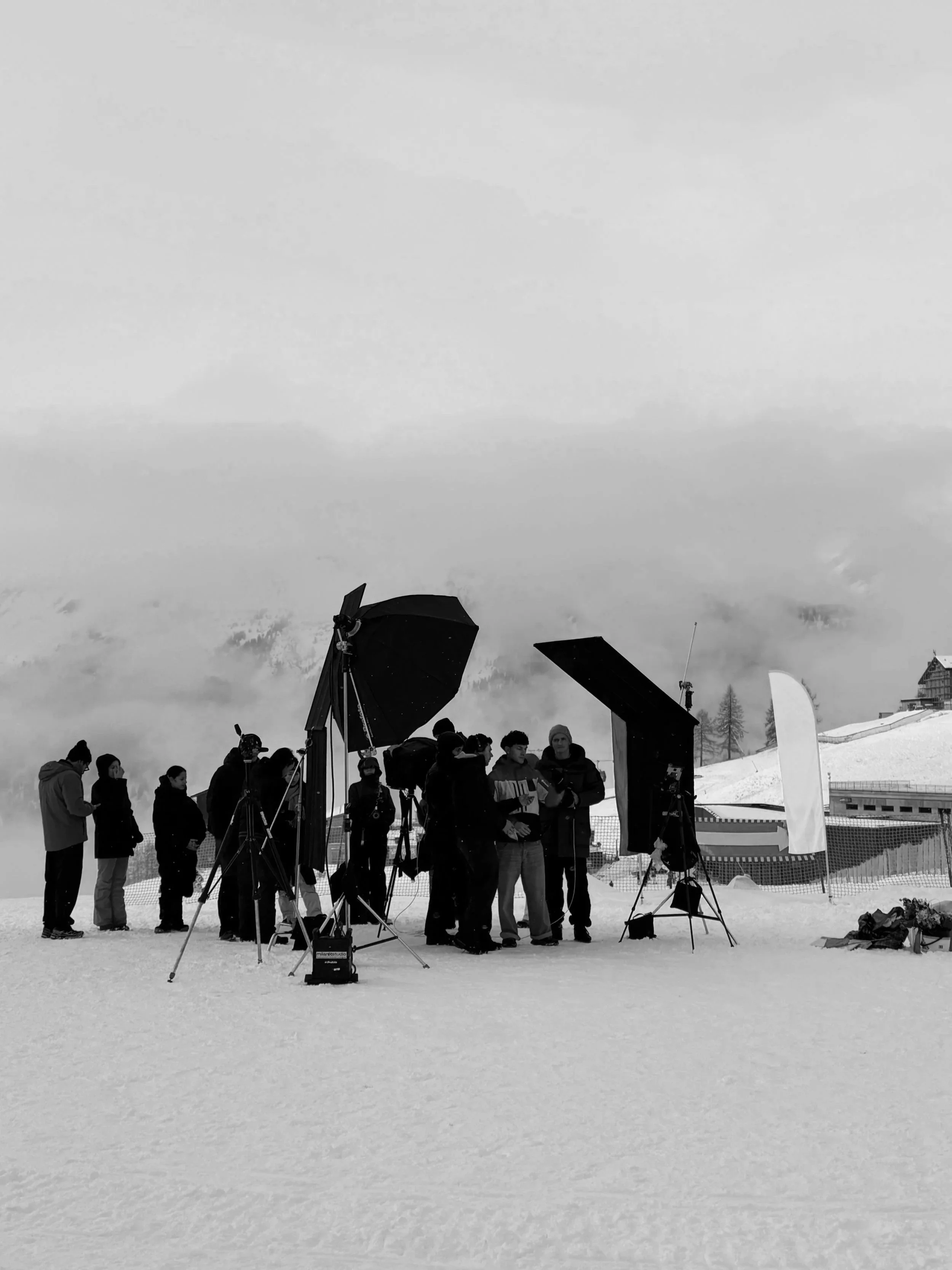 Group of people gathered on snow-covered ground with photography equipment, including large lights and umbrellas, in a snowy mountainous area.