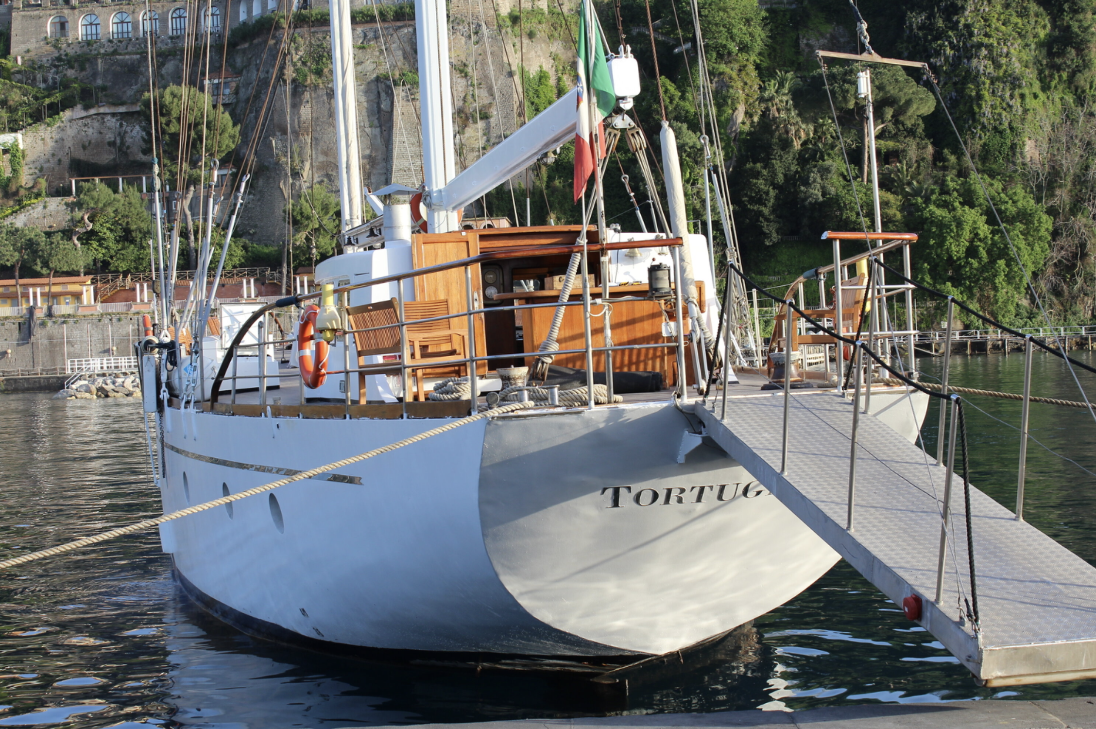 A sailboat named Turtuga docked at the marina with a gangway connecting it to the pier. The boat has wooden accents, a flag, and several masts and ropes. There's green foliage and hillside buildings in the background.