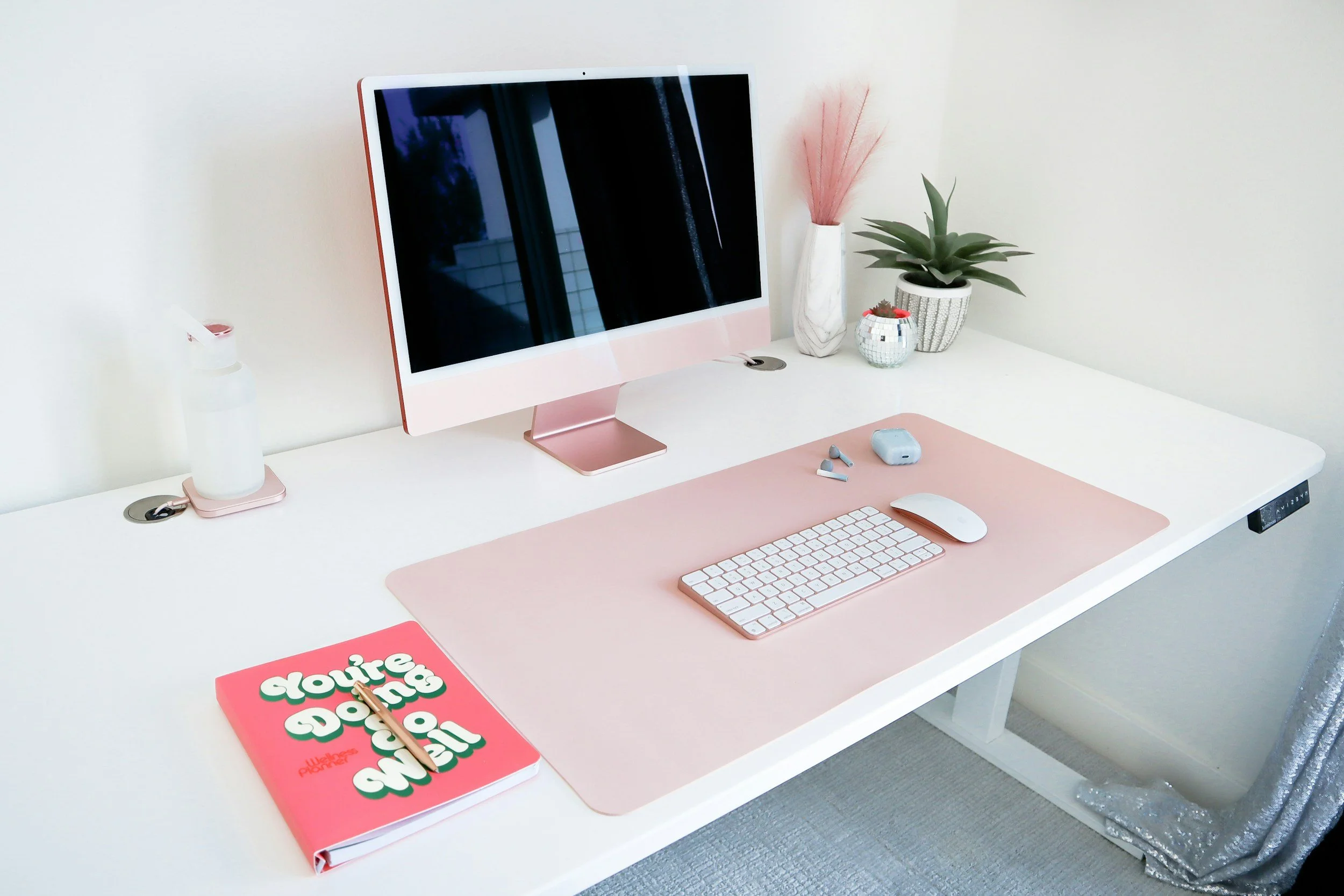 A white desk with a pink and white computer monitor, a pink keyboard, a white mouse, wireless earbuds, a blue case, and a pink notebook with colorful text. The desk also has a bottle of hand sanitizer, a pink decorative vase with pink pampas grass, two potted plants, and a small decorative orb. The desk area is tidy and decorated with pink and white accents.
