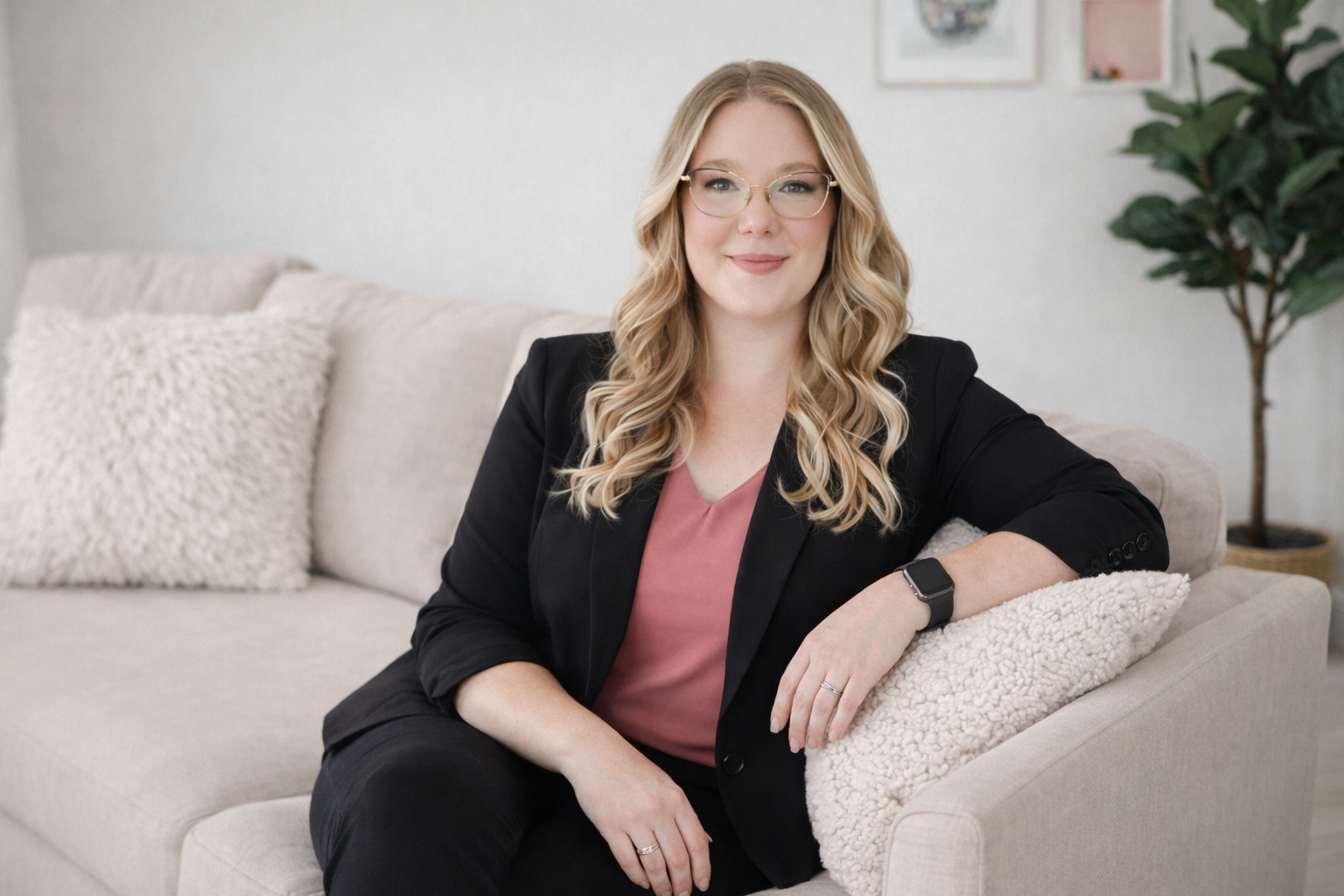 A woman with blonde wavy hair, glasses, wearing a black blazer, pink top, black pants, and a smartwatch, sitting on a light-colored sofa with textured pillows, in a bright room with wall art and a plant in the background.