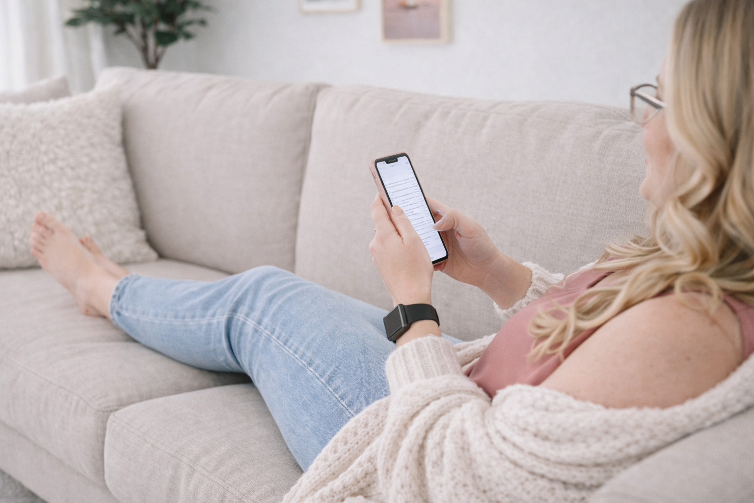 A woman relaxing on a beige sofa, holding and looking at her smartphone, with her legs extended on the sofa, wearing light blue jeans, pink sleeveless top, cream knit cardigan, and glasses, in a cozy living room with a potted plant and framed pictures in the background.
