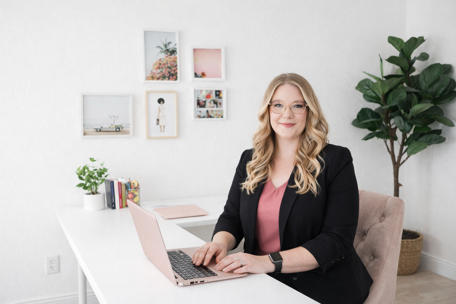 A woman with blonde curly hair and glasses sitting at a white desk with a pink laptop, wearing a black blazer and pink top, in an office with white walls, framed pictures, green plants, and office supplies.