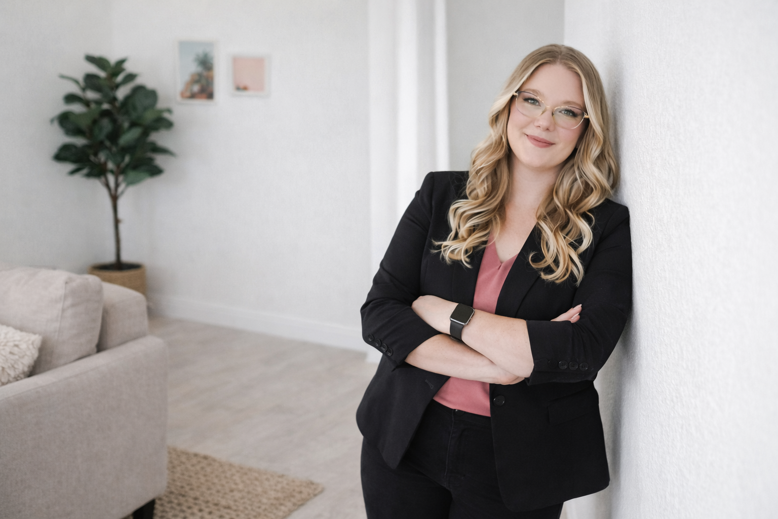 A woman with blonde curly hair, glasses, and a pink blouse, standing with crossed arms and smiling while leaning against a white wall in a bright, modern living room.