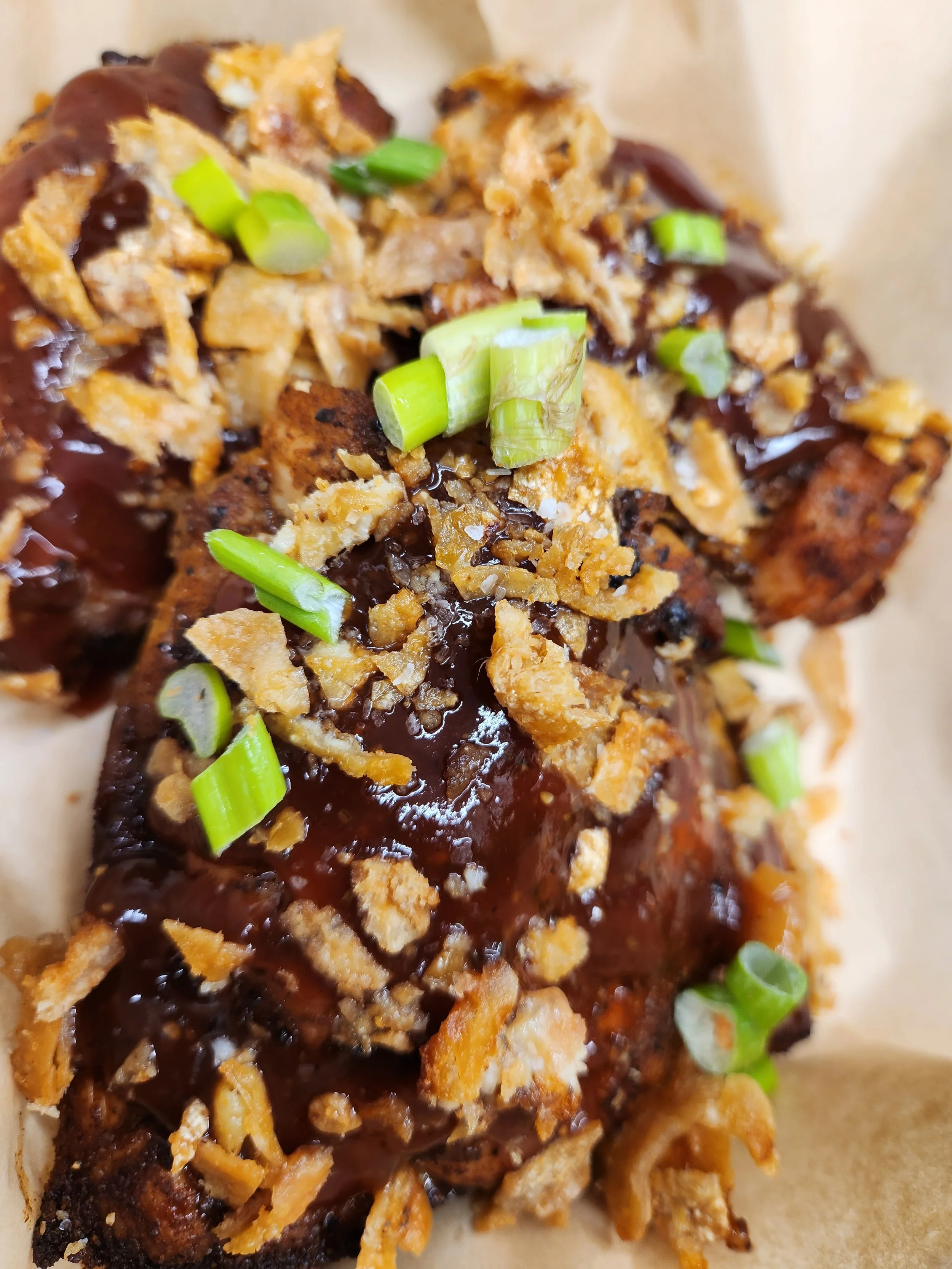 Close-up of a fried food item topped with chopped green onions, crispy fried onions, and a dark sauce, served on a piece of paper or parchment.