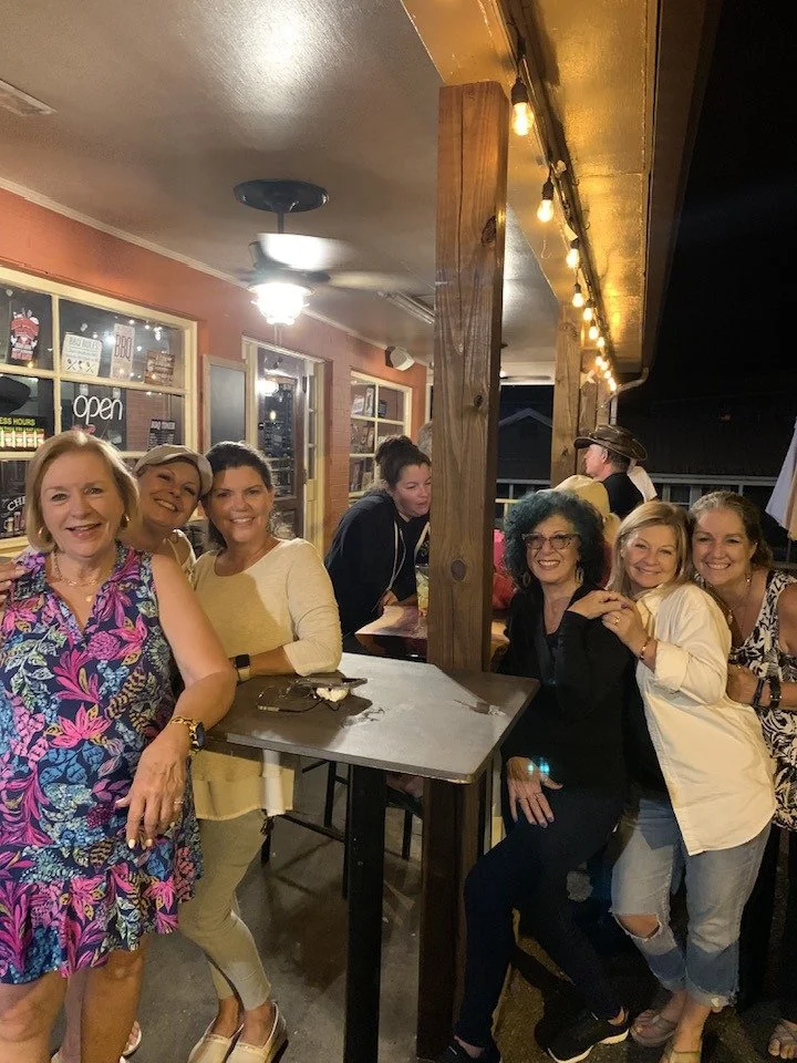 Group of women smiling and posing for a photo inside a restaurant or bar, with some sitting and some standing, dressed casually, with warm lighting and a rustic interior.