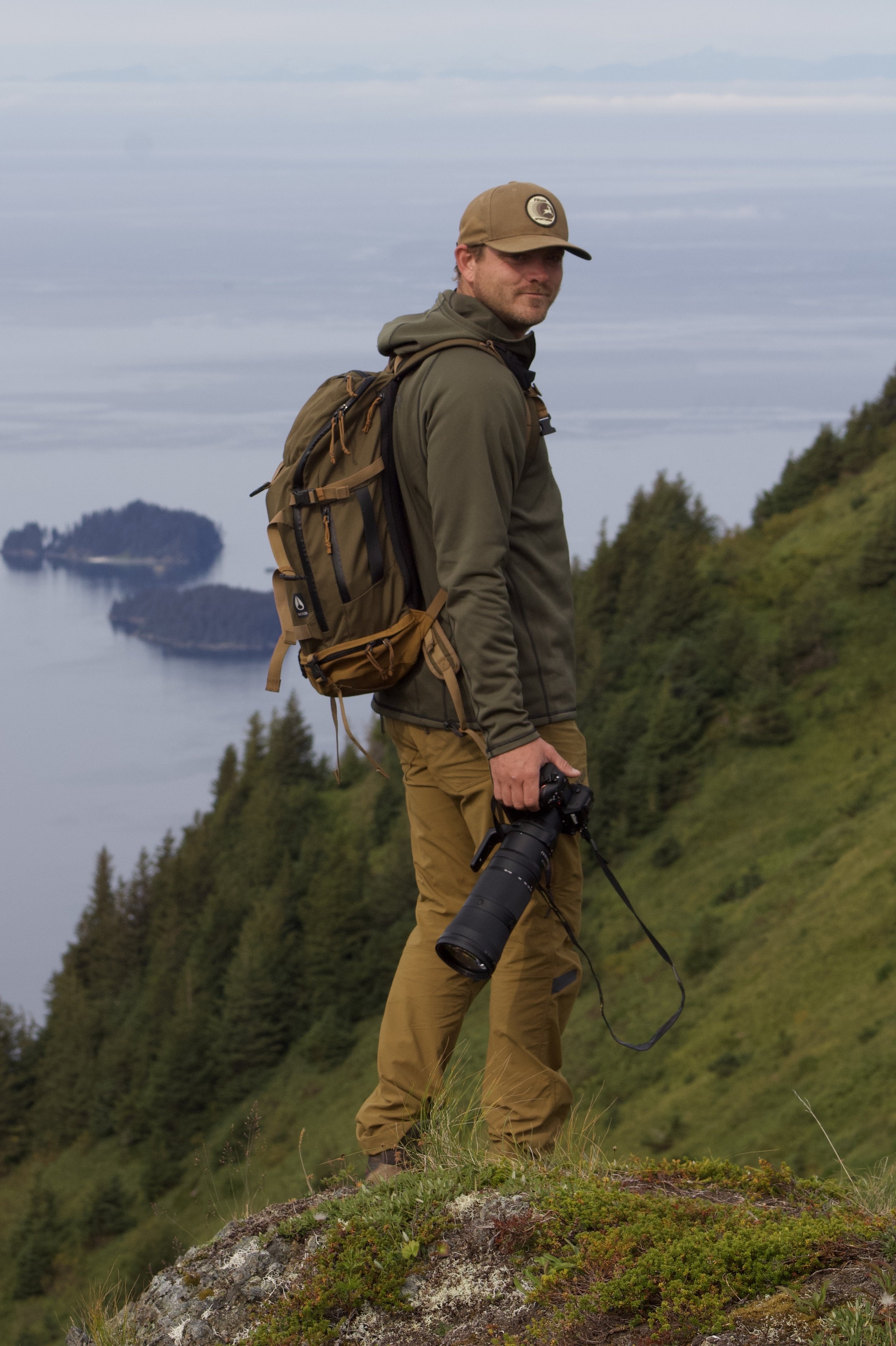A man standing on a grassy hilltop holding a camera with a long telephoto lens, dressed in outdoor hiking gear, with a backpack, overlooking a scenic landscape of green hills, water, and small islands under a cloudy sky.