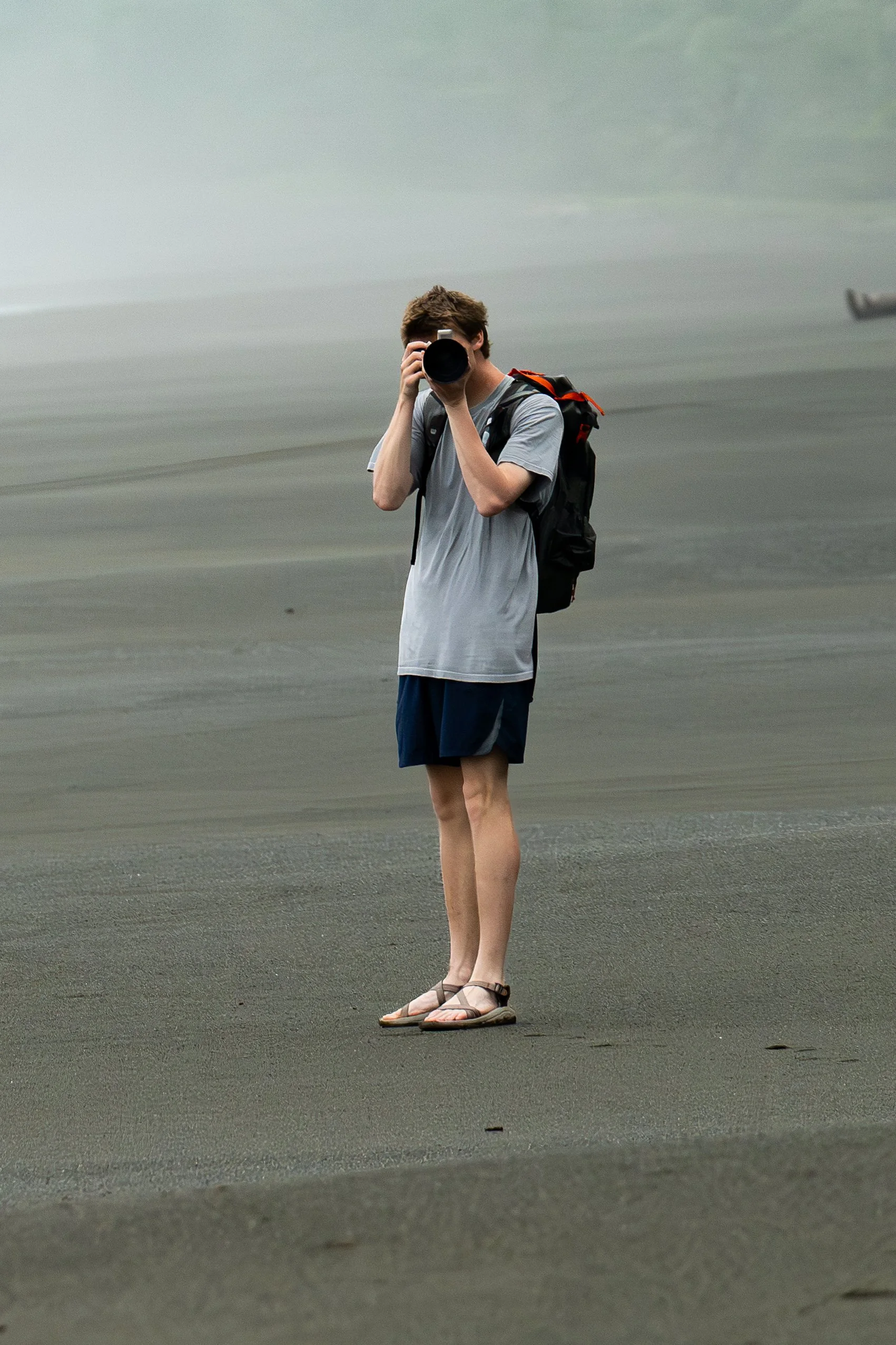 Person standing on sandy beach taking a photo with a camera, wearing a gray t-shirt, shorts, sandals, and carrying a backpack.
