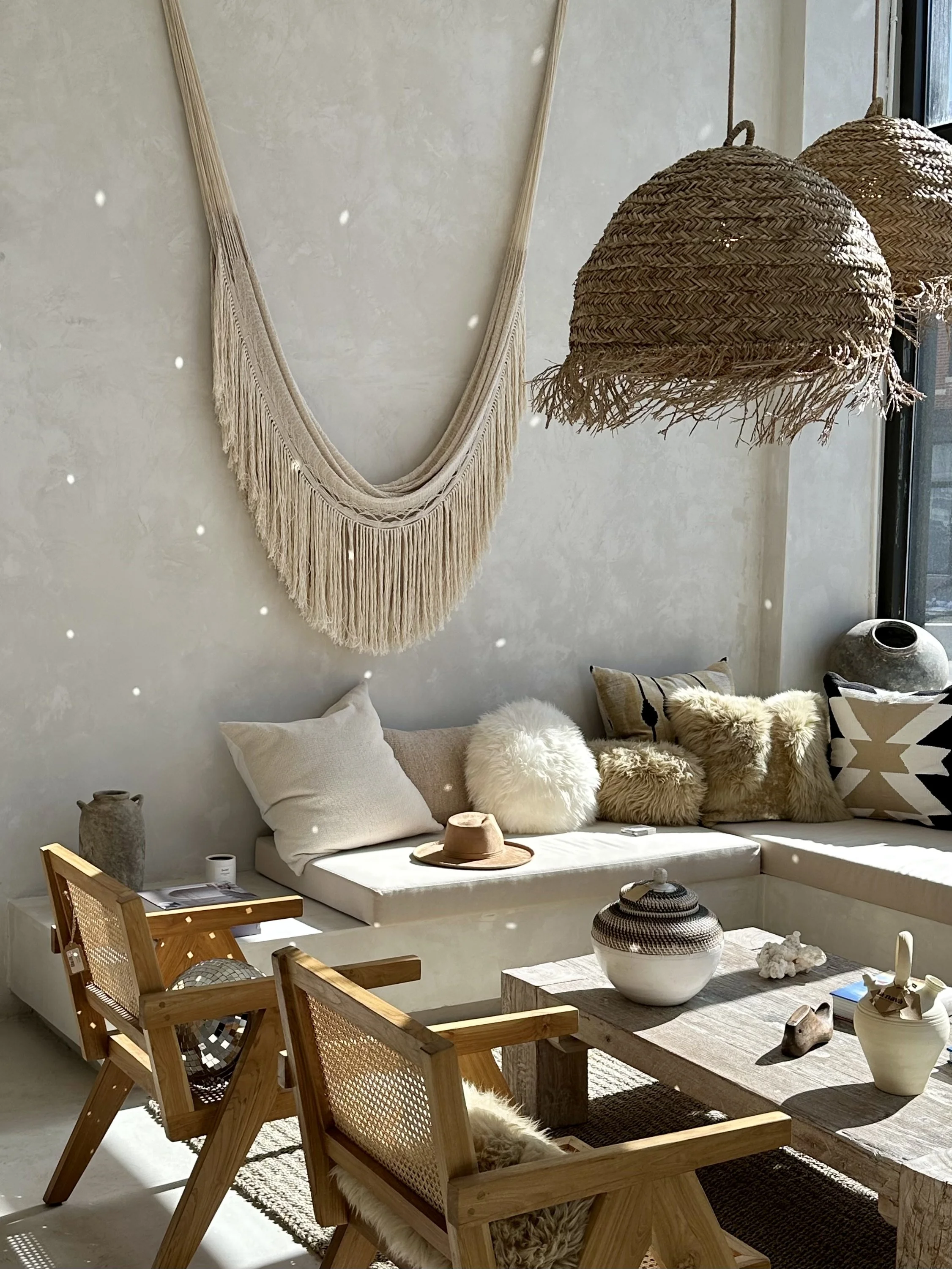 A cozy living room with bohemian decor features a white built-in sofa with various fluffy and patterned pillows. A beige hat rests on the sofa. There is a rustic wooden coffee table with decorative vases and small objects. Two wooden armchairs with fur throws are placed in front of the table. The wall behind has a large, cream-colored macramé wall hanging, and two woven pendant lights hang from the ceiling. Sunlight streams in through a nearby window.