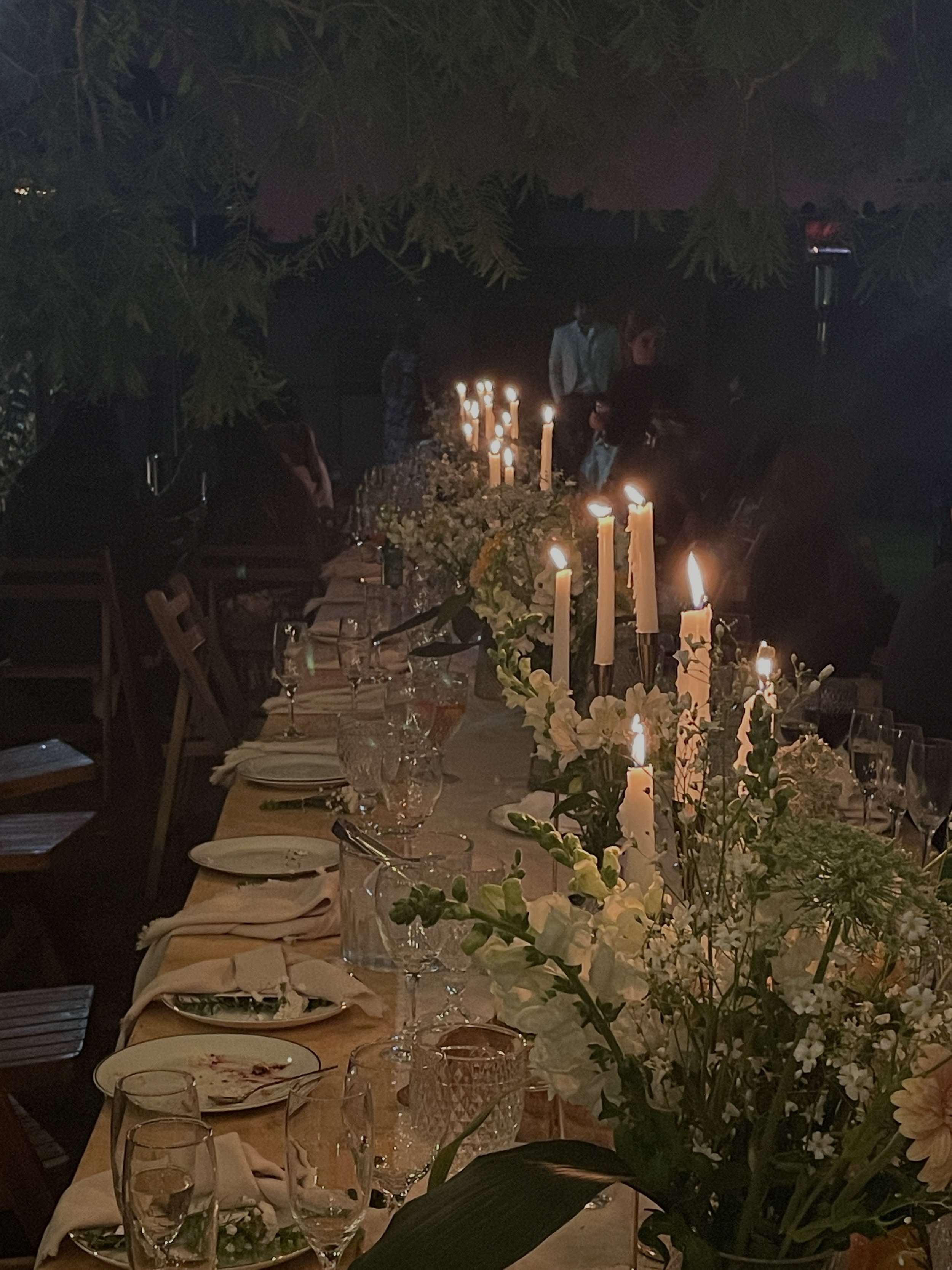 A long dining table decorated with white flowers and lit candles in a dimly lit setting, possibly for a wedding or special event. The table is set with glasses, plates, and napkins, and there are people in the background, some standing and some seated.