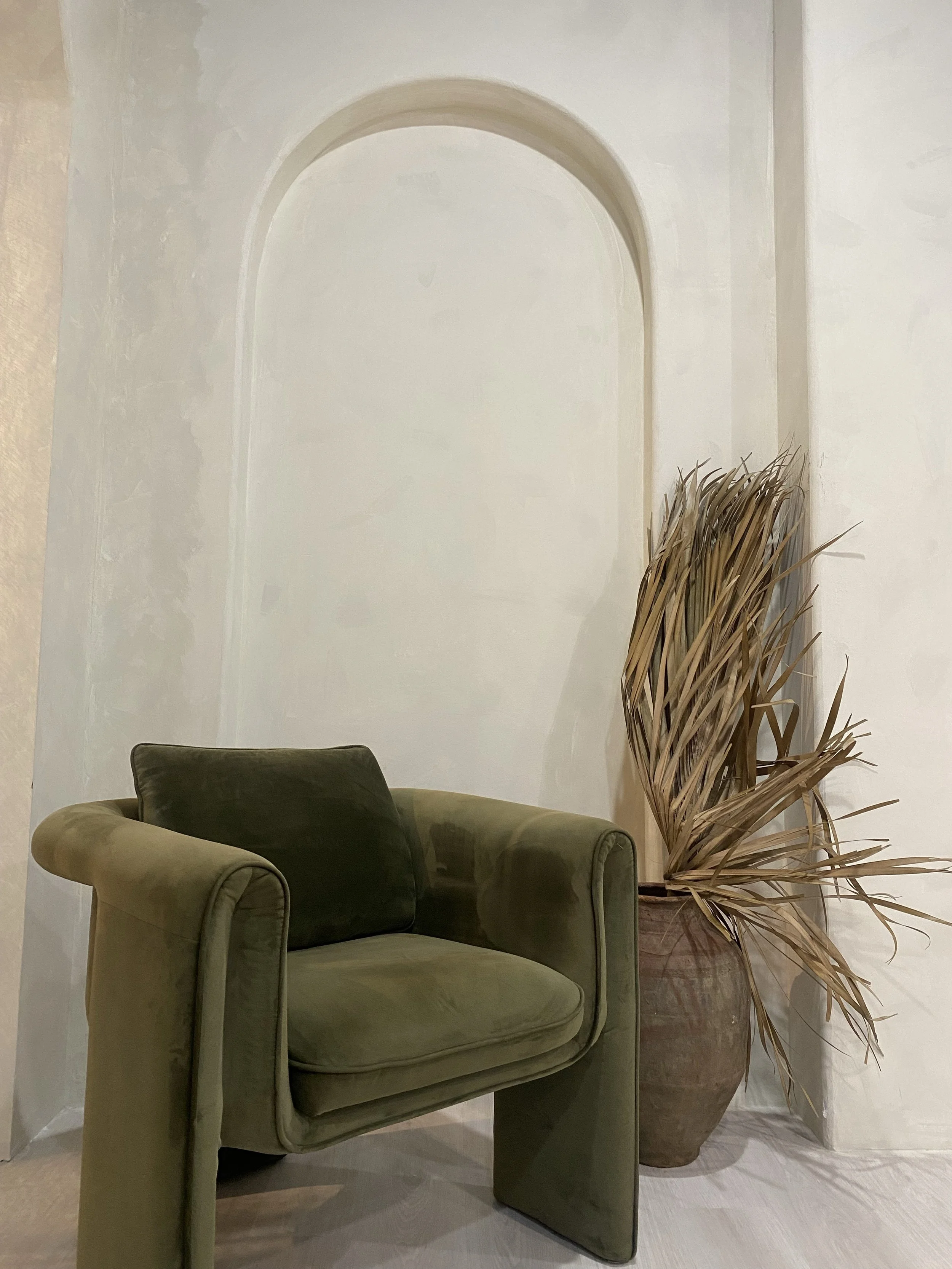 A green velvet armchair with a dark mulberry cushion, placed next to a large terracotta pot filled with dried, beige-colored plants, against a white wall with an arched alcove.