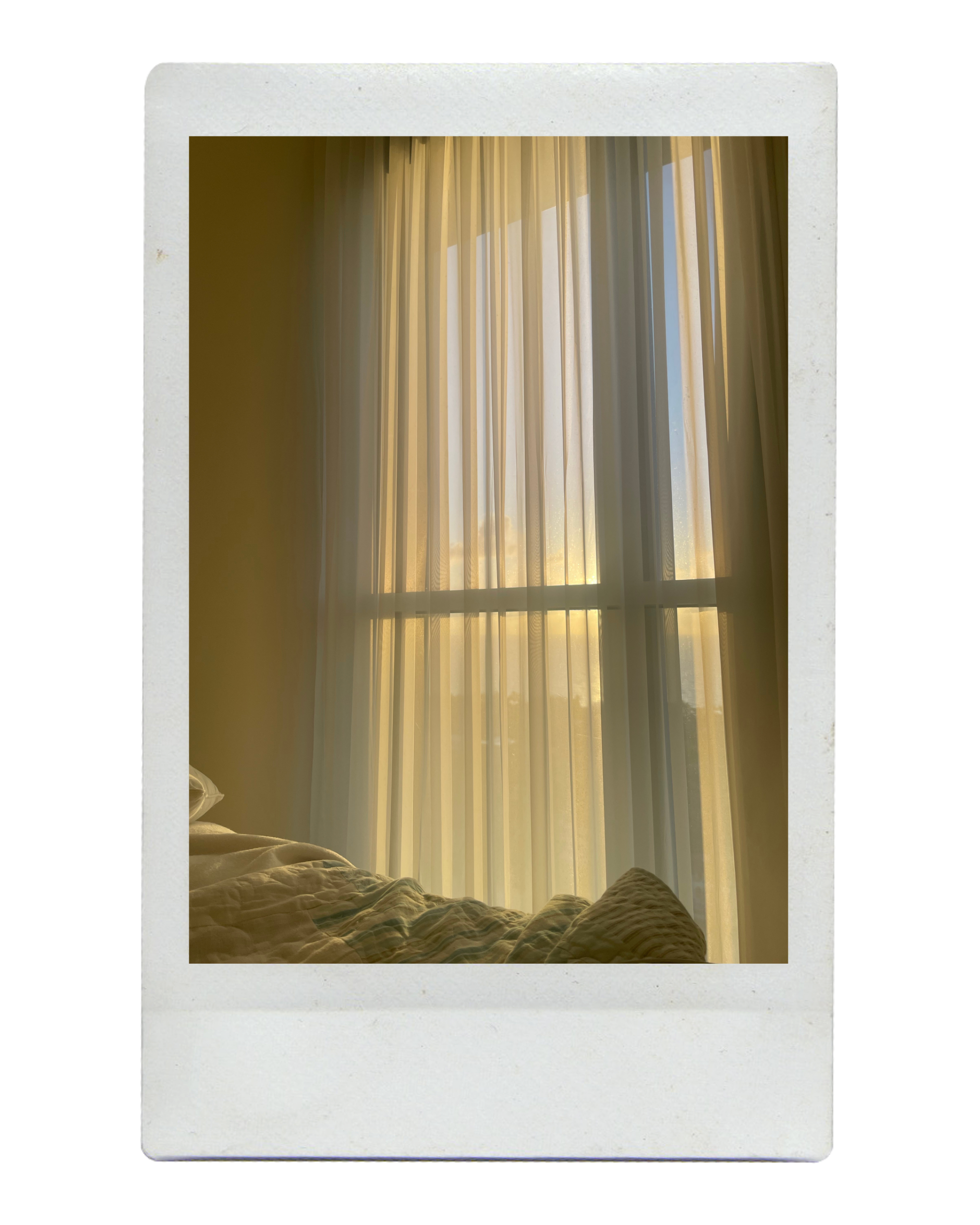View of a window with closed curtains and sunlight shining through, with part of a bed in the lower left corner.