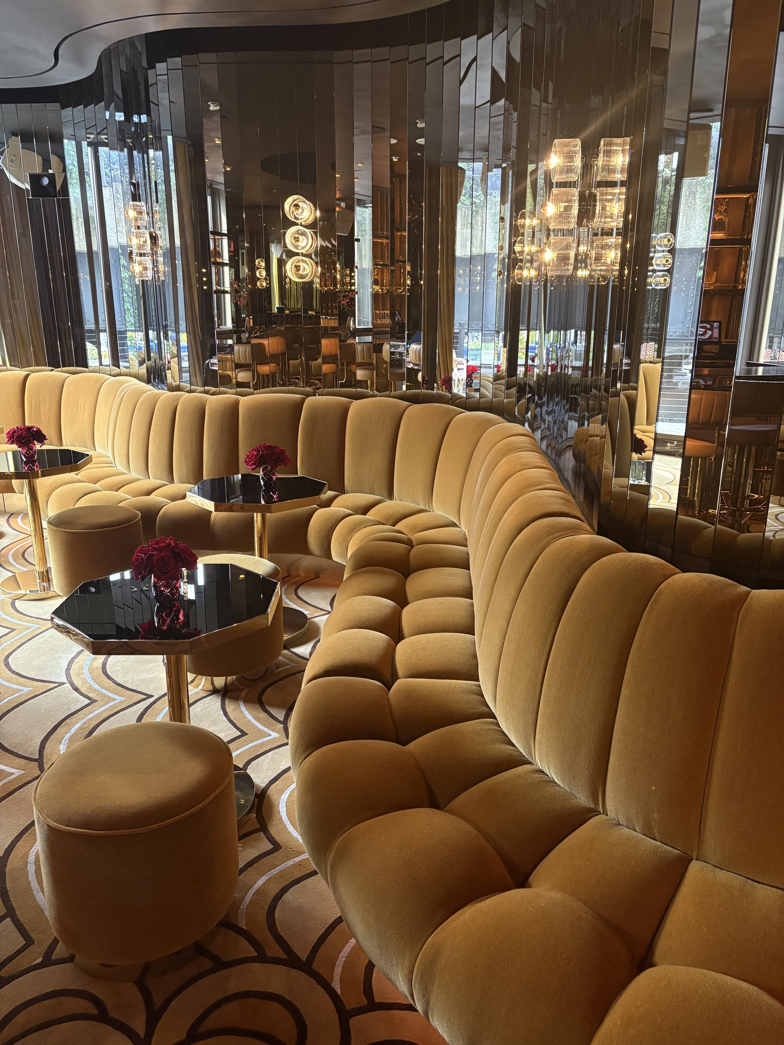 Interior of a luxurious hotel lounge or restaurant with a curved mustard-yellow sofa, small black topped tables with red roses, plush carpet with intricate patterns, and reflective modern lighting fixtures and mirrors.