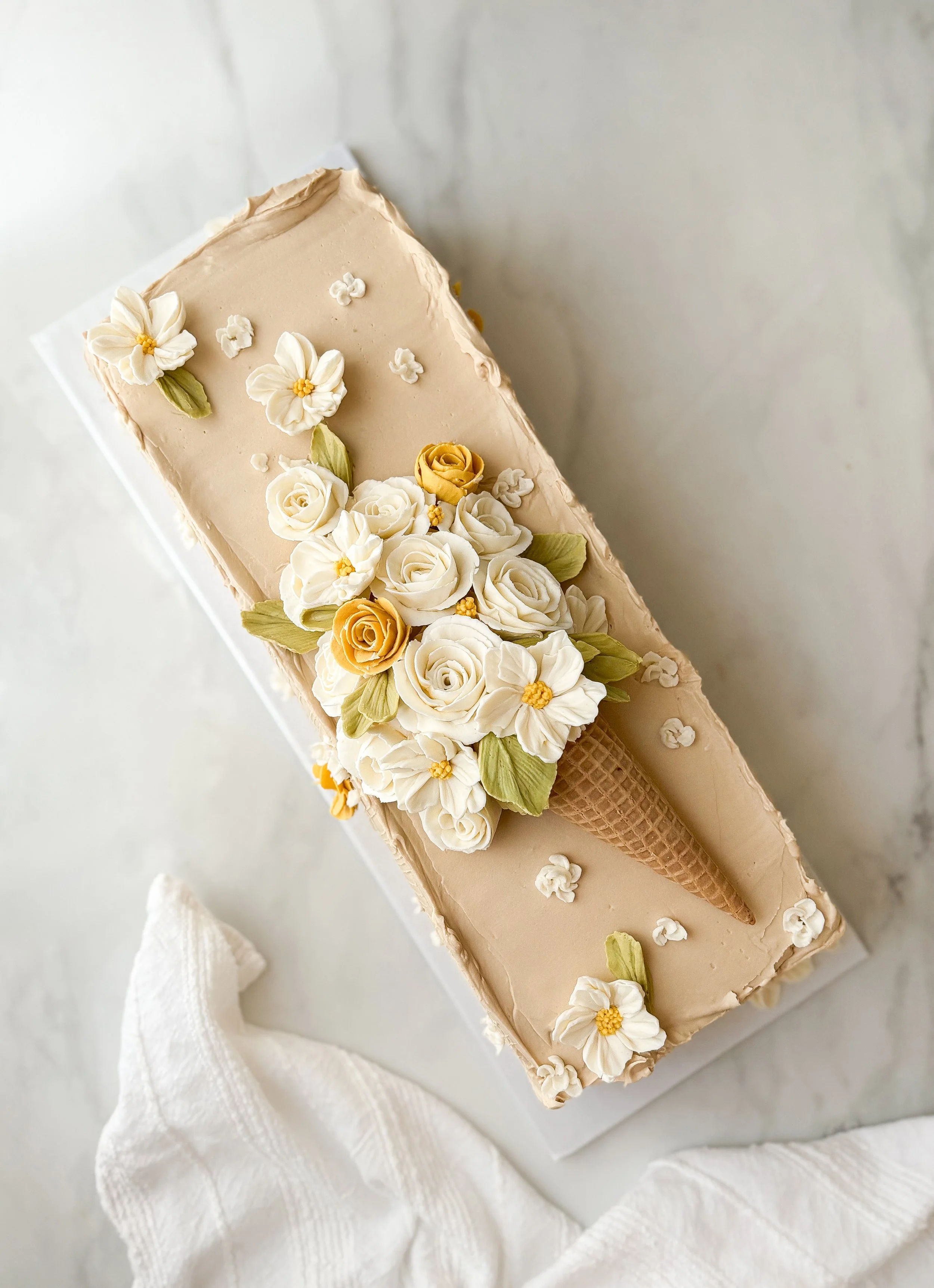 A rectangular cake decorated with white and yellow flowers, with an ice cream cone made of icing on top, placed on a white surface with a white cloth nearby.