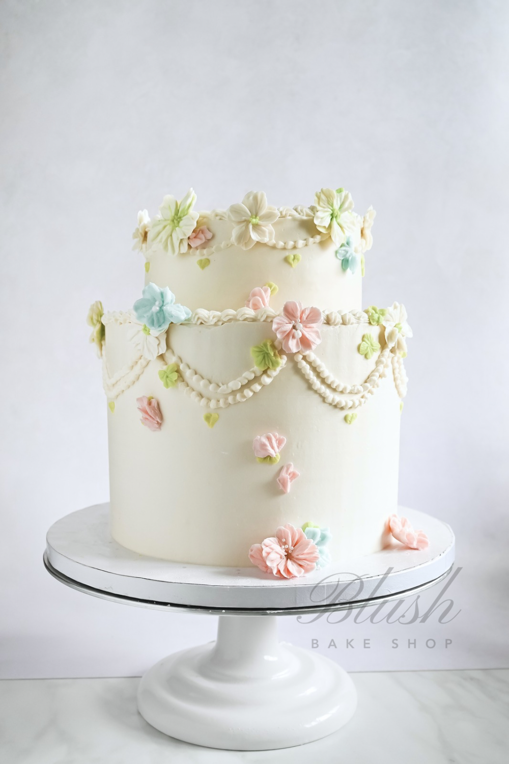 Two-tiered white wedding cake decorated with pastel flowers and pearl-like icing garlands on a white cake stand.
