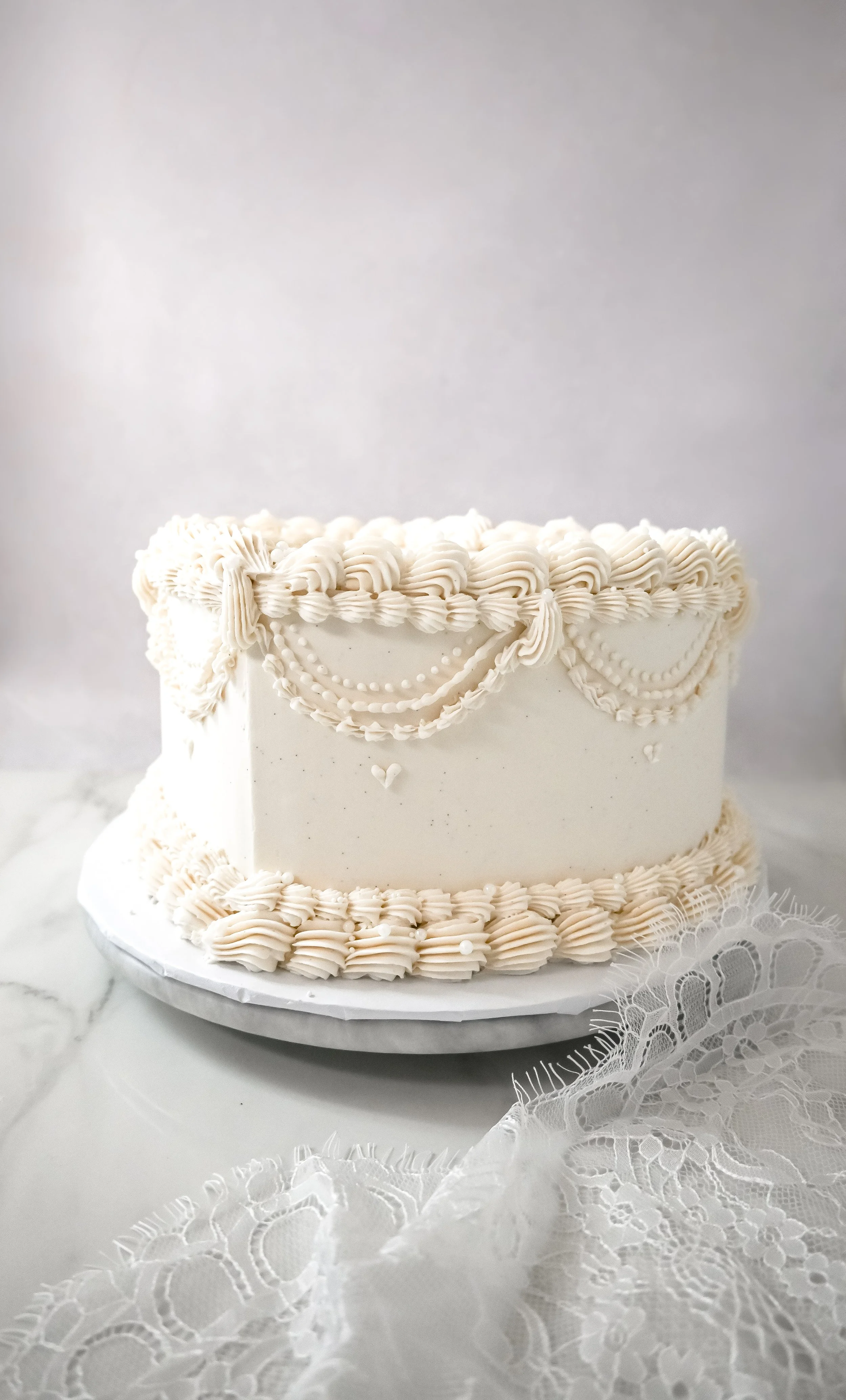 White wedding cake with ornate piped icing decoration on top and around the edges, placed on a white marble surface with lace fabric in front.