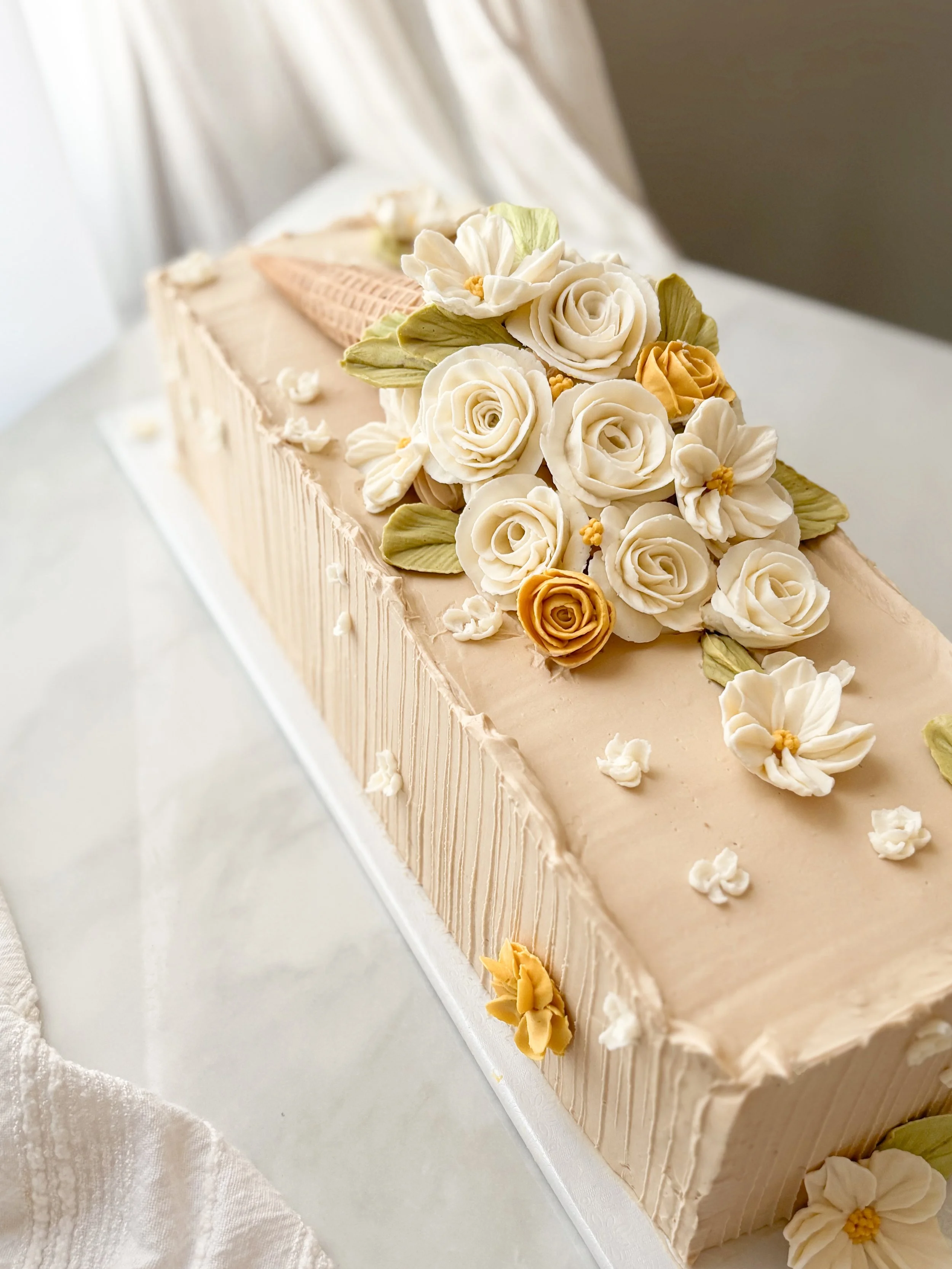 A rectangular wedding cake with beige frosting, decorated with white and yellow fondant roses, white flowers, and green leaves.