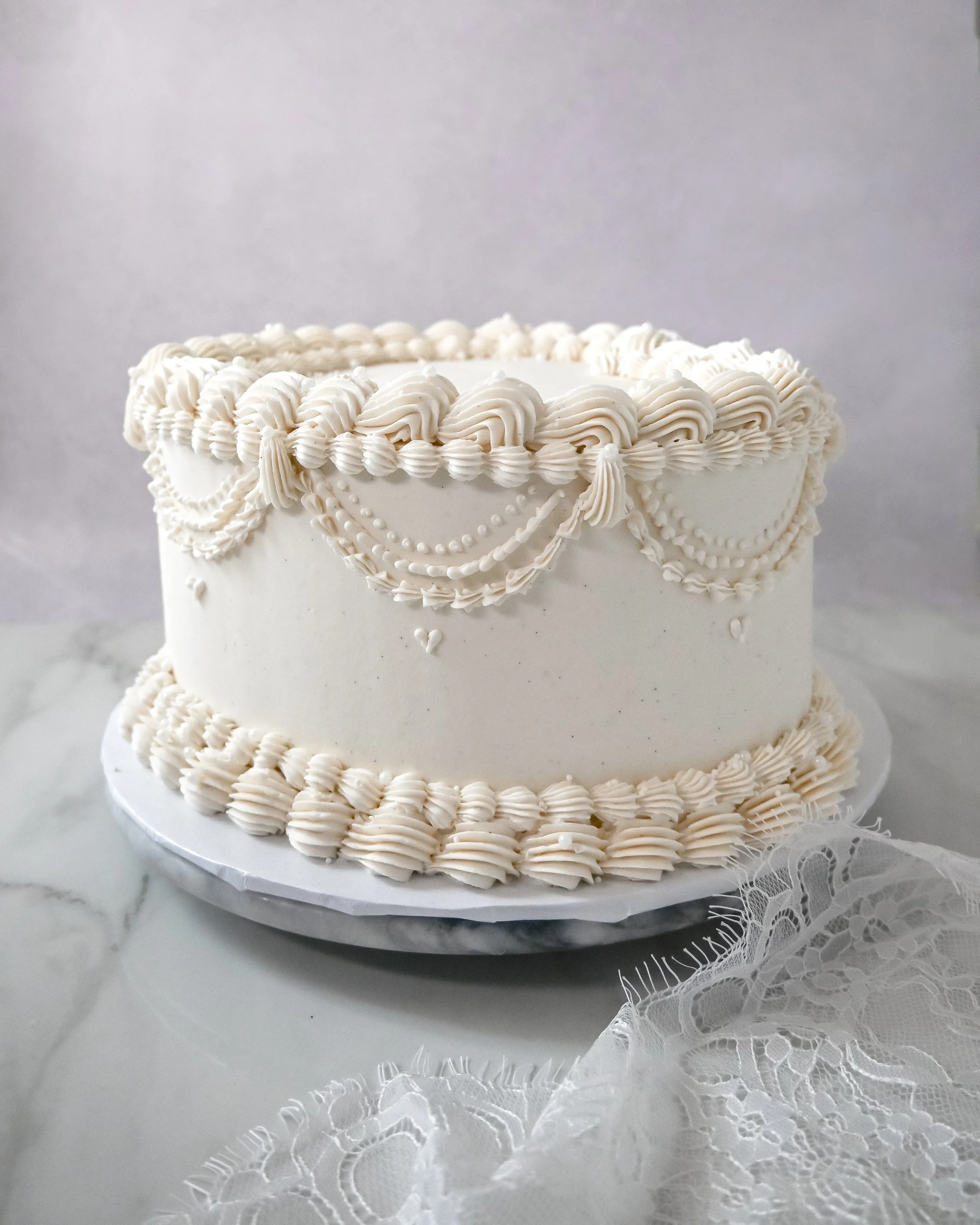A white vintage wedding cake with decorative piped borders and icing details, placed on a marble surface with lace fabric nearby.