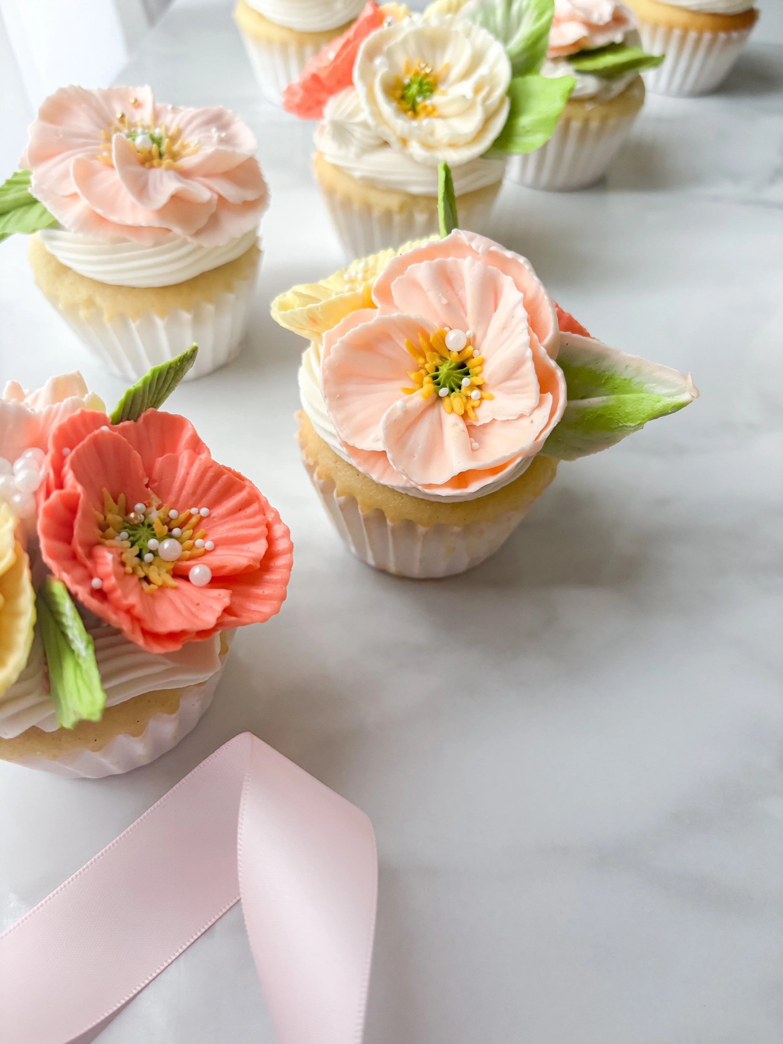 Decorative cupcakes with pastel-colored flower and leaf icing decorations on a white surface, with a light pink ribbon in the foreground.