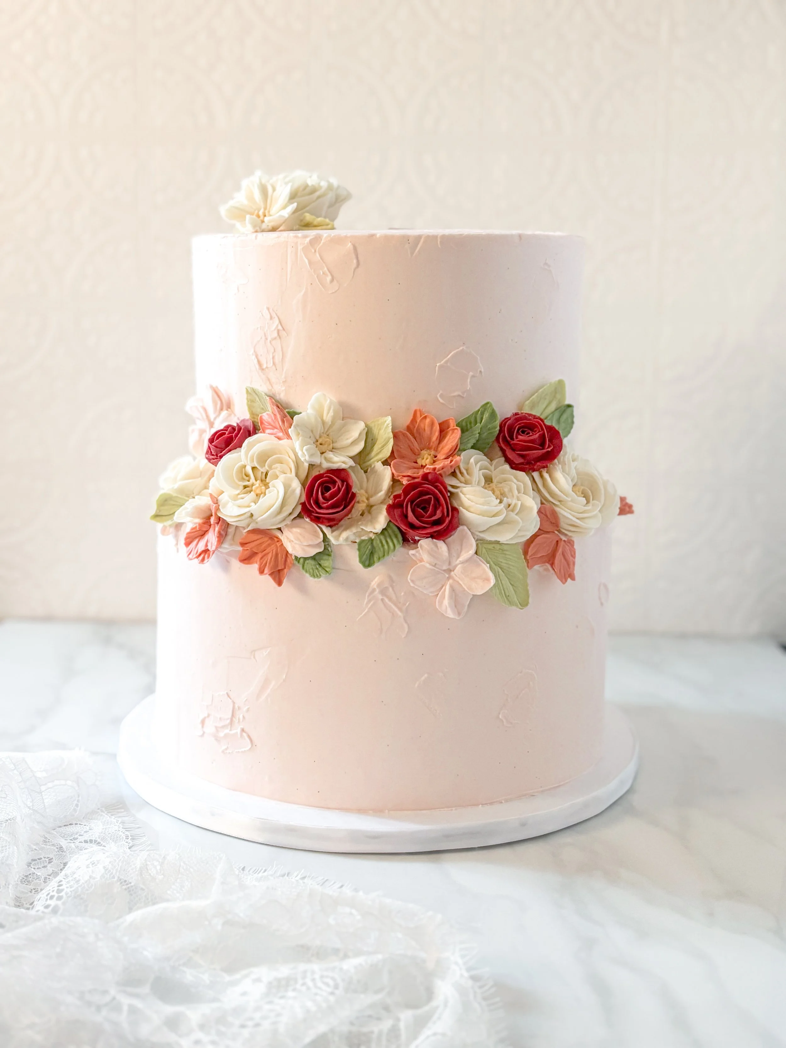 Pink two-tiered wedding cake decorated with white and red flowers and green leaves, placed on a white marble cake stand.