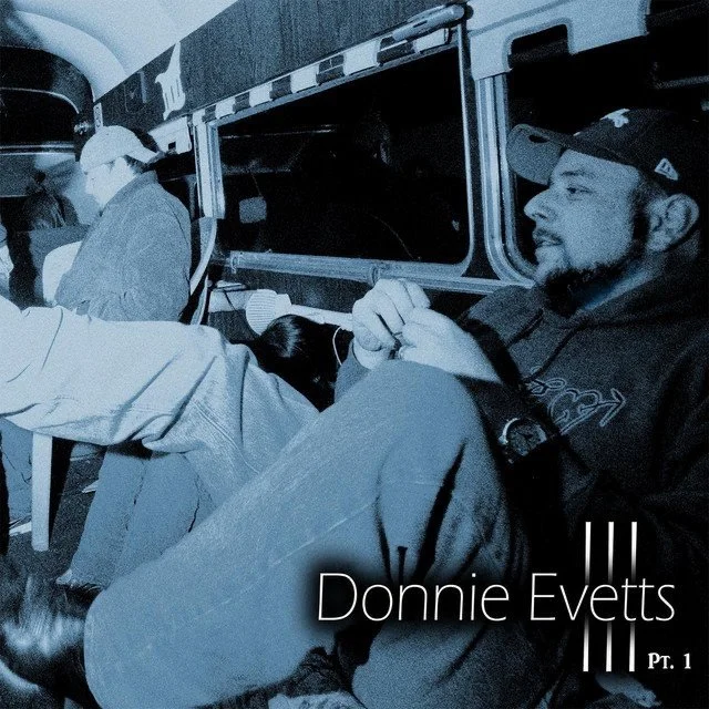 A man with a beard and a cap sitting on a bus, with his hands clasped, looking out the window. Other passengers are visible in the background.