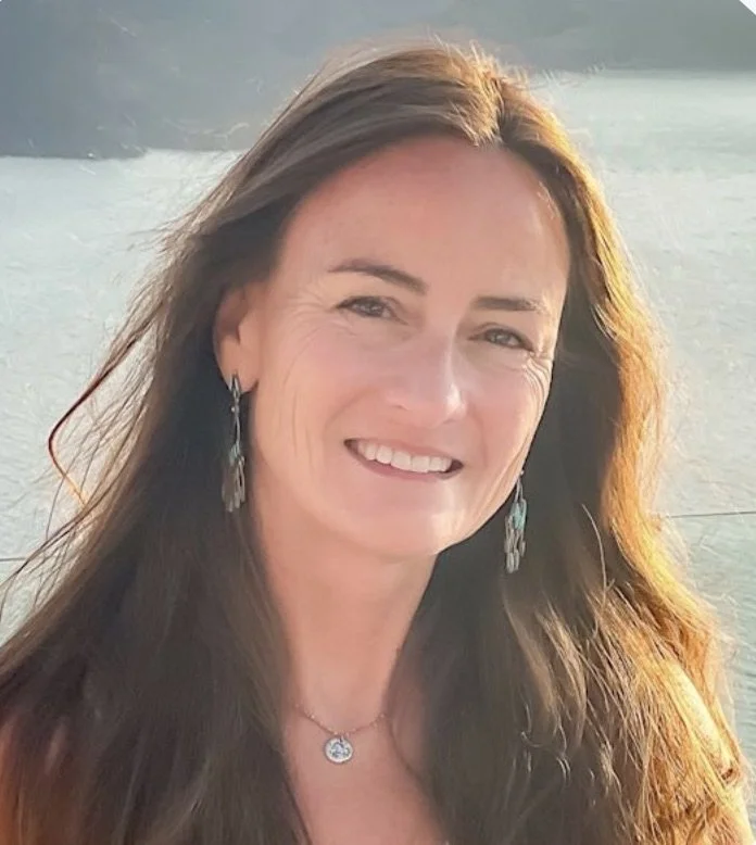 A woman with long brown hair smiling with water and faint mountains in the background.