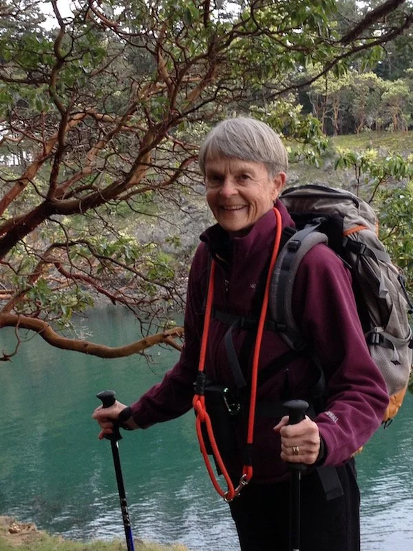 An older woman with short gray hair smiling outdoors, holding trekking poles, wearing a backpack, near a body of water and surrounded by trees.
