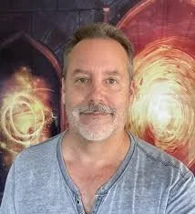 A middle-aged man with a beard smiling in front of a colorful abstract background with swirling light patterns.